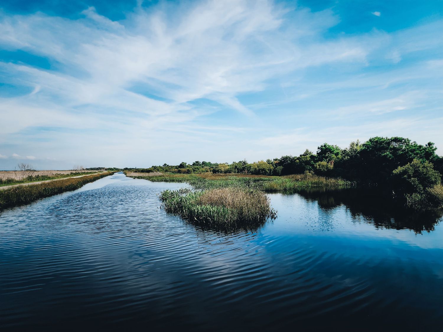 Tranquil waterway with marsh grass and blue skies reflecting on calm water surface.