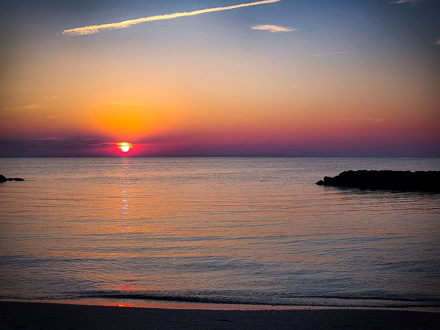 Vibrant orange sunset over calm ocean waters with silhouetted rocky coastline.