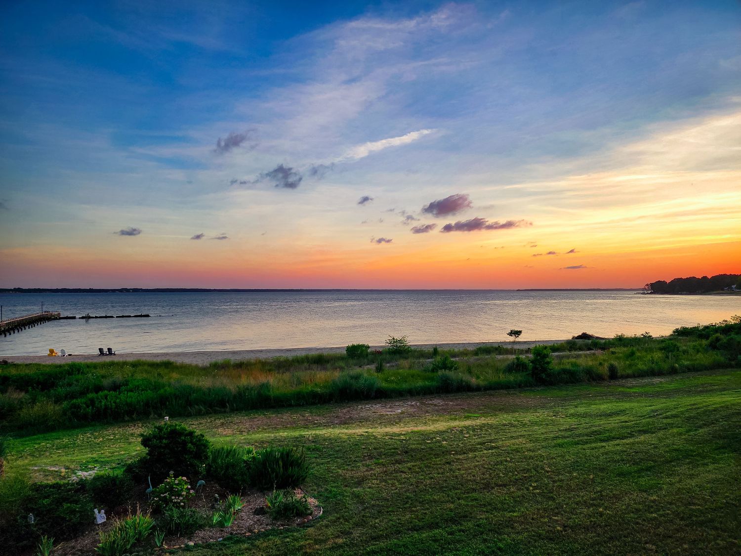 Peaceful beach sunset with orange sky reflecting on calm waters and green coastal grounds.