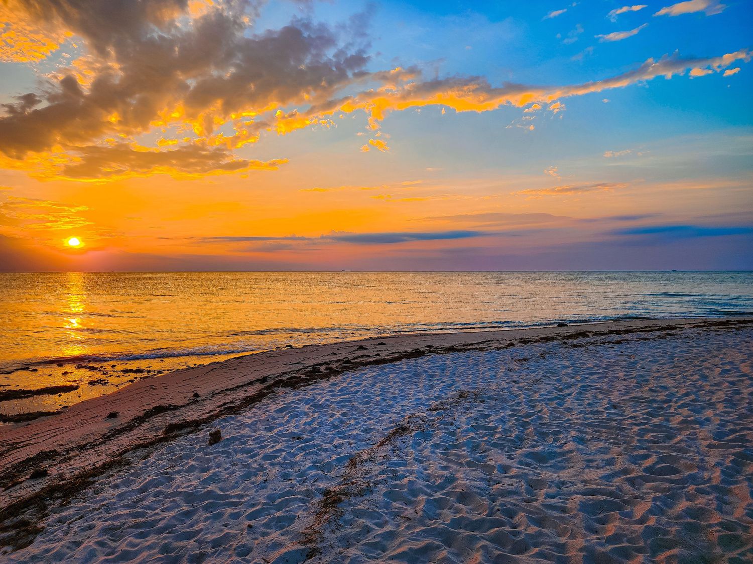 Golden sunset illuminates beach shoreline with orange clouds reflecting on calm ocean waters.