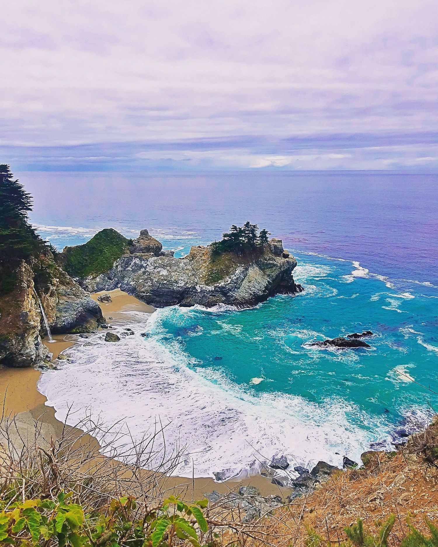 Turquoise waves crash against rocky cliffs and sandy beach at McWay Falls in Big Sur, California.