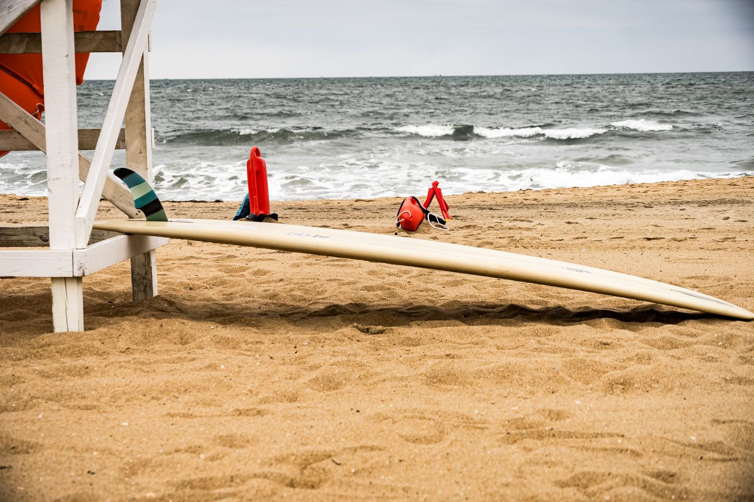 A red and white lifeguard rescue surfboard stands upright on a sandy beach near the ocean waves.