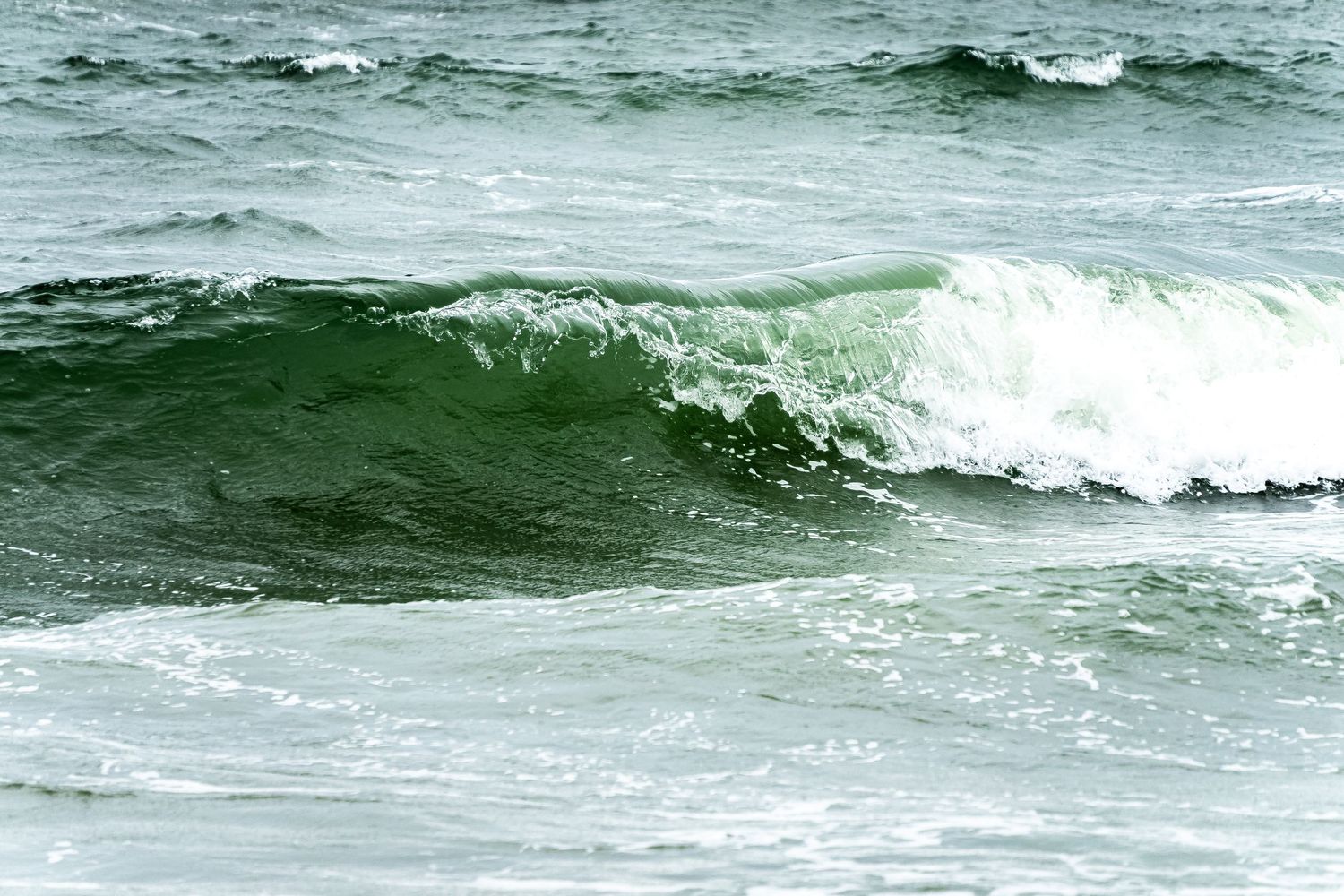 Rolling ocean waves with white foam crests break in a sequence across turquoise green waters on a cloudy day.