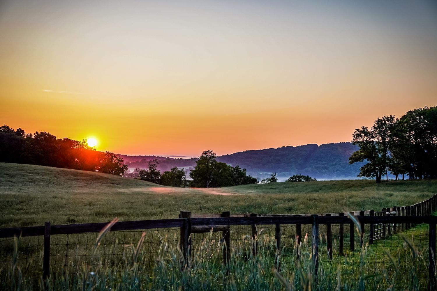 Scenic sunset over rolling farmland with wooden fence in foreground and silhouetted trees against orange sky.