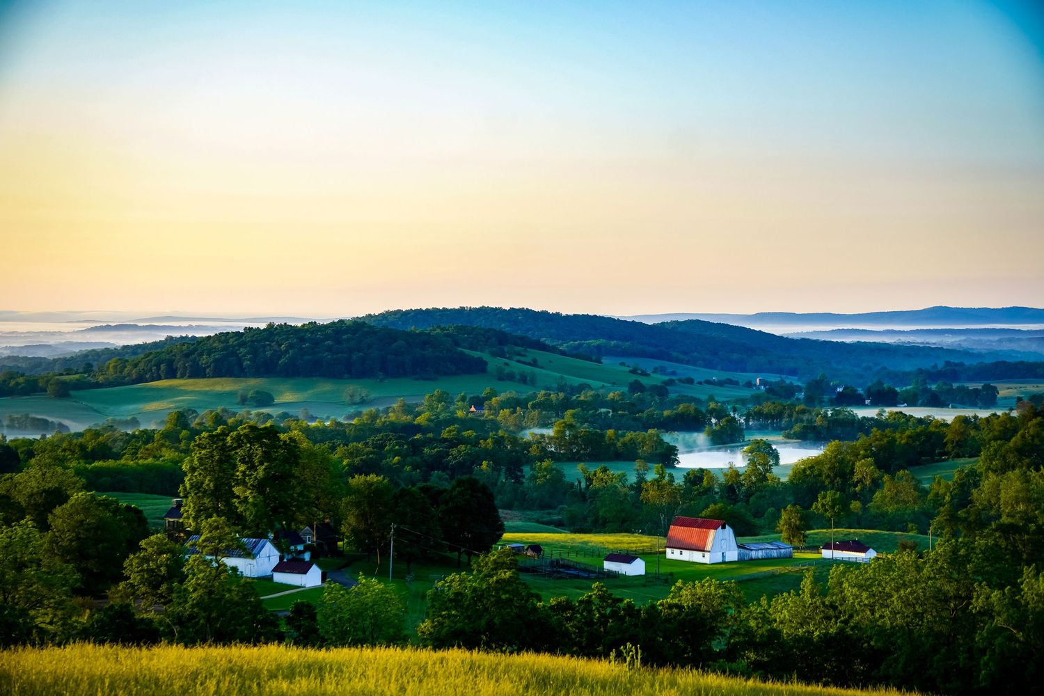 Misty morning view of rural valley with white barns and farmhouses nestled among green hills.