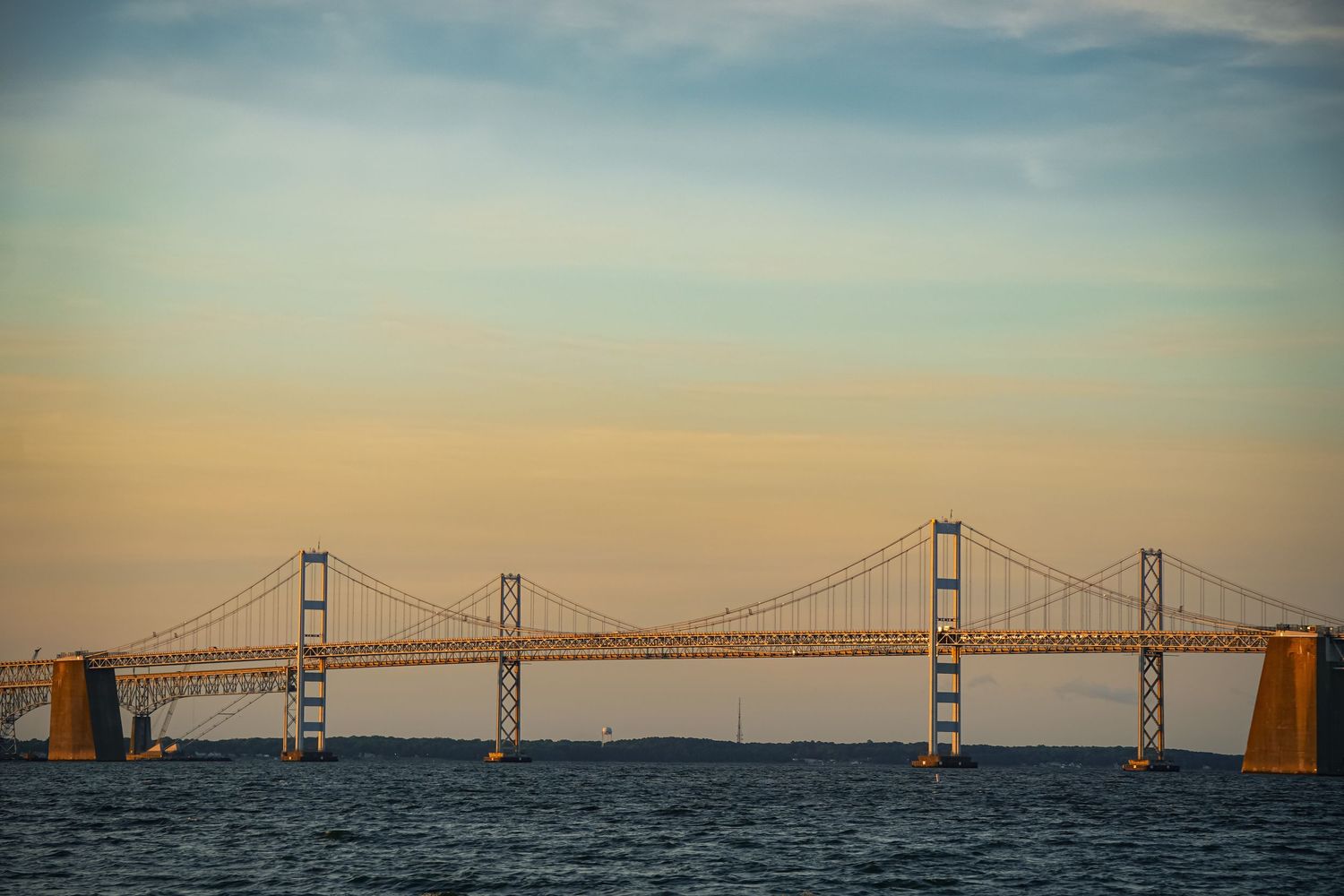 The Chesapeake Bay Bridge spans across calm waters during sunset with a golden-orange sky.