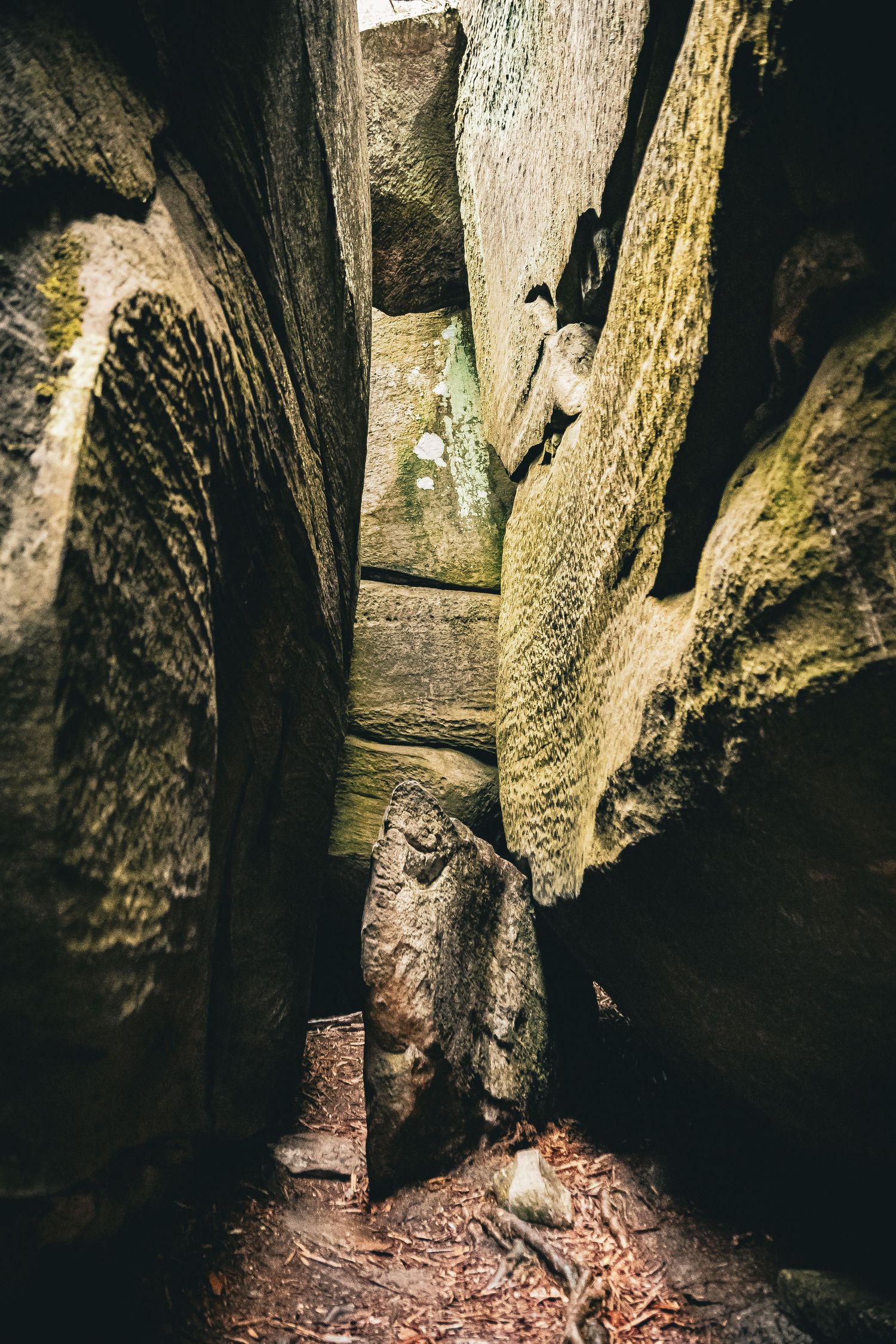 Narrow passage between tall moss-covered rock formations in a dark cave system with natural lighting from above.