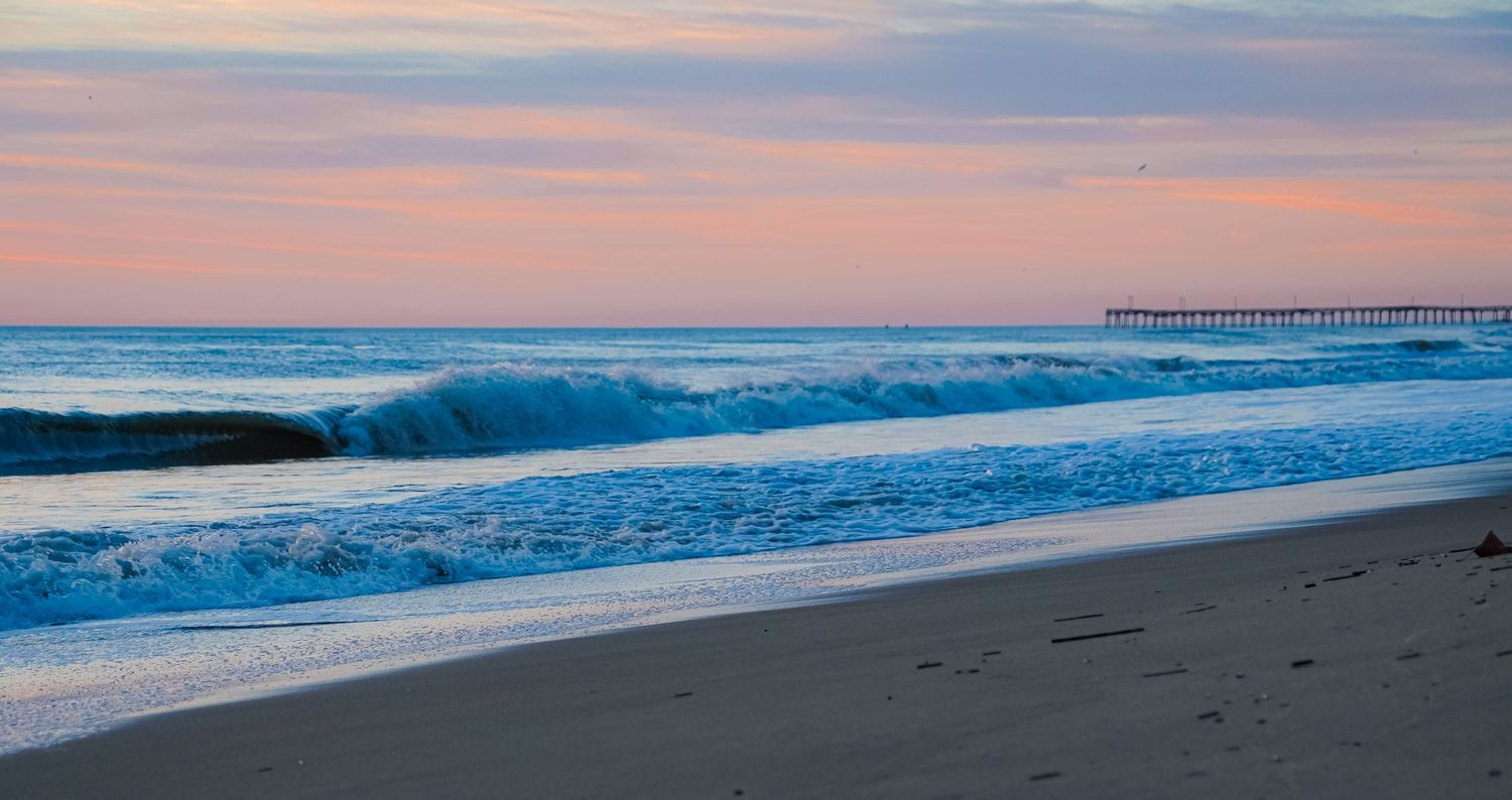 Ocean waves roll onto sandy beach at sunset with pier visible in distance.