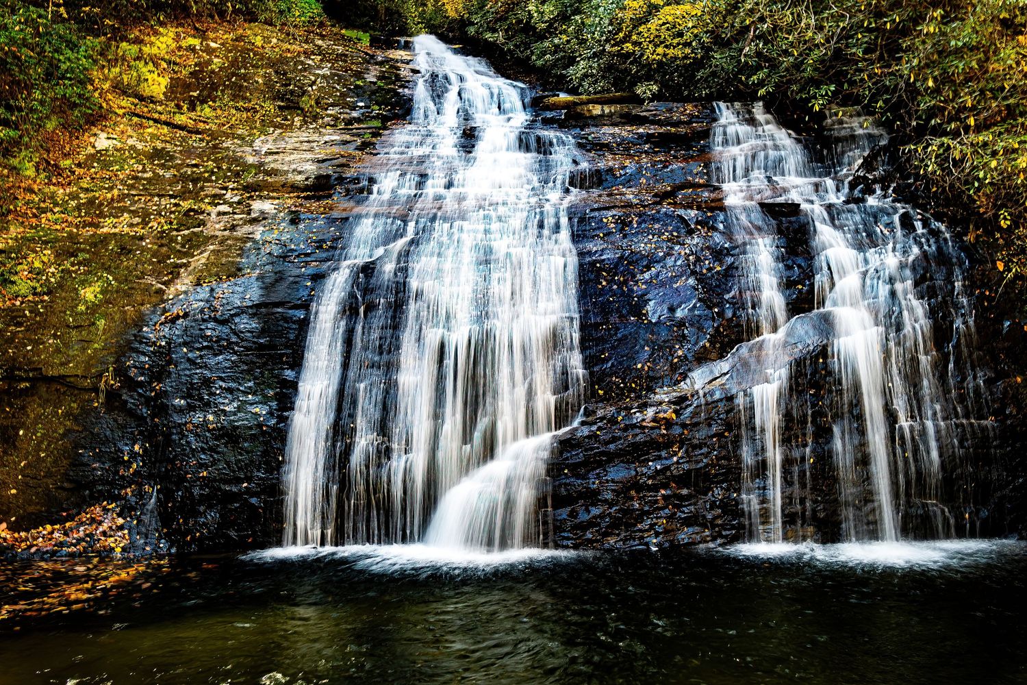 Twin waterfalls flow dramatically over dark rocky cliffs into a serene forest pool.