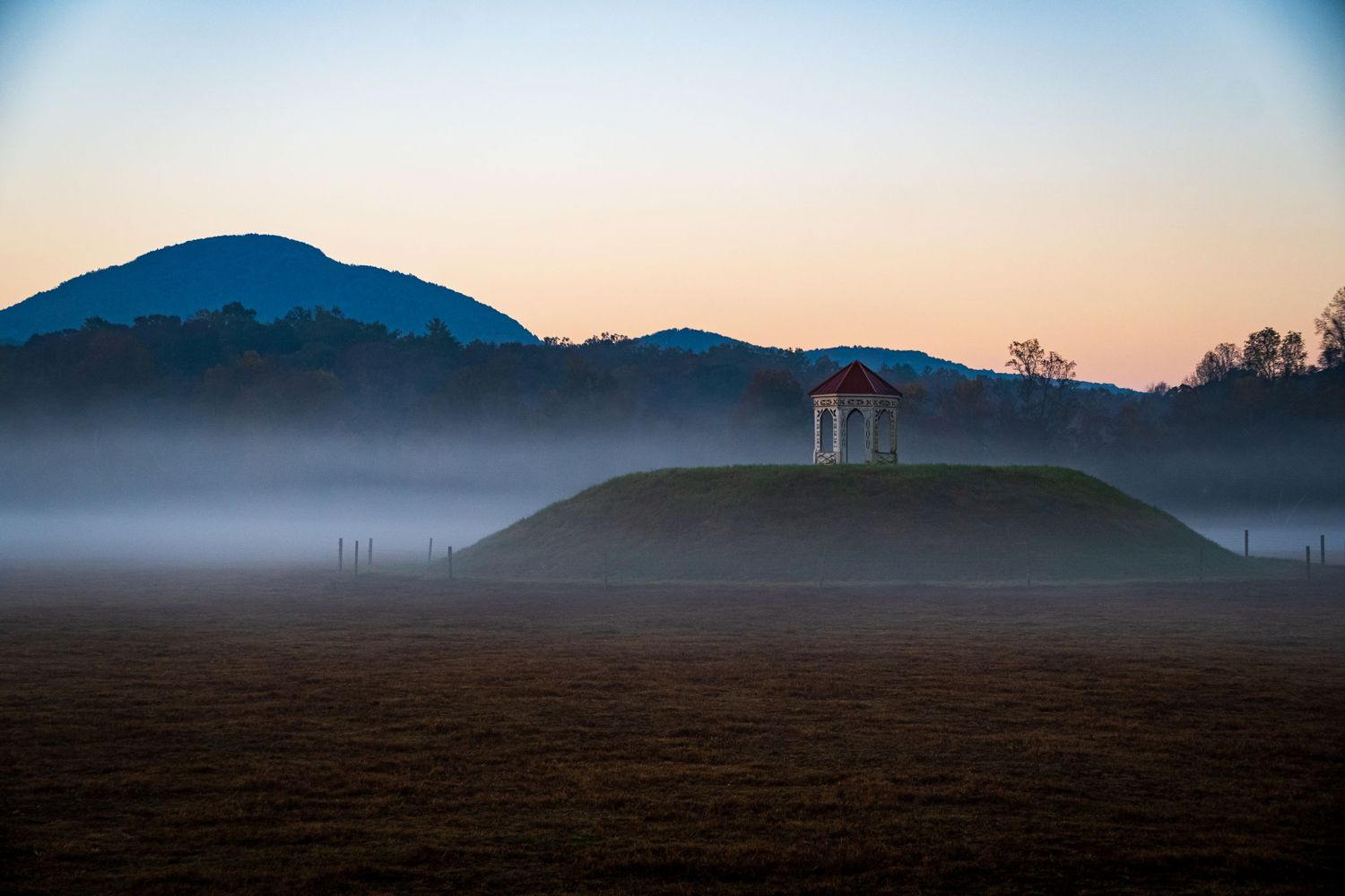 Small gazebo on hilltop silhouetted against mountains and morning mist.