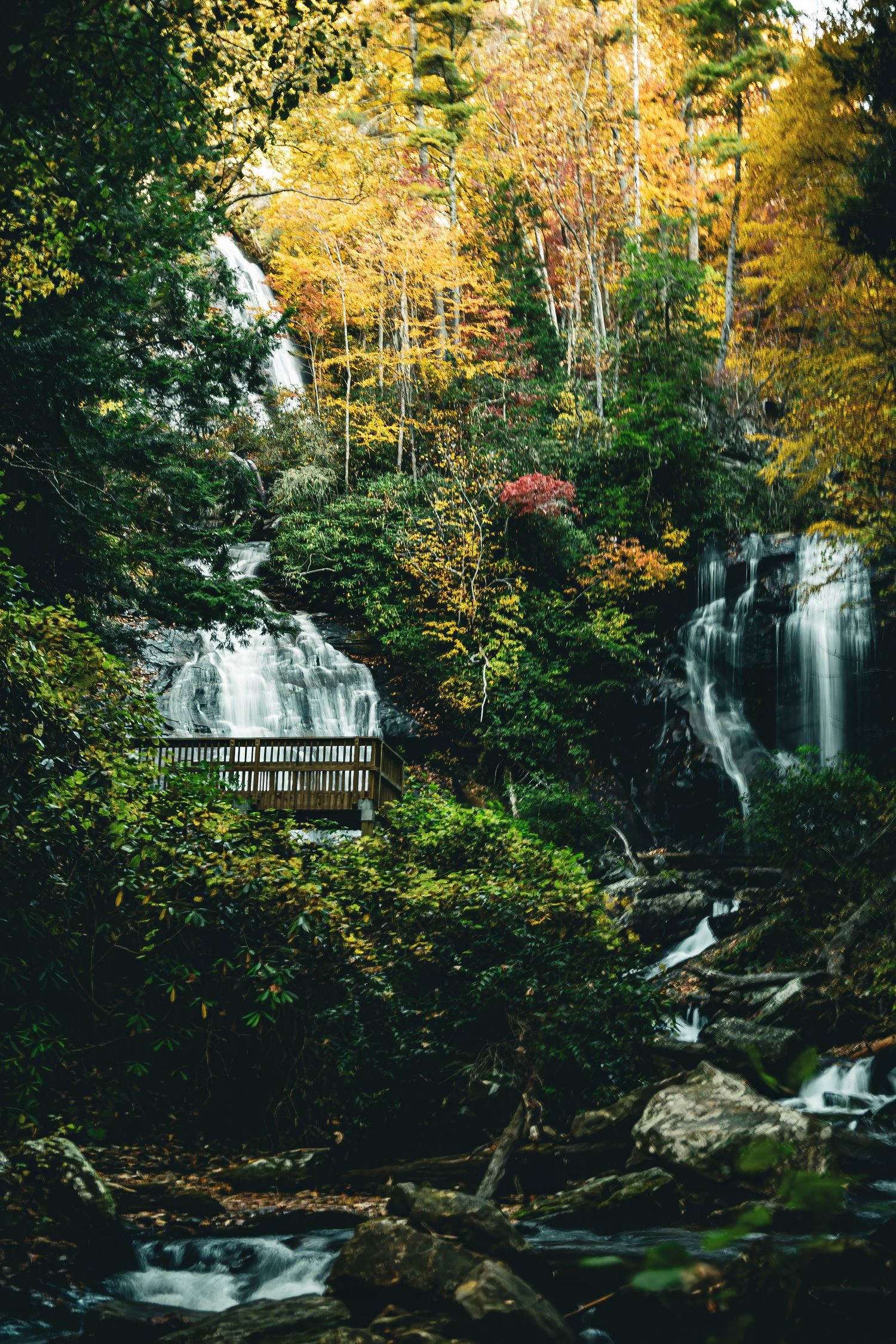 Twin waterfalls flowing past wooden observation deck surrounded by autumn trees in mountain forest.