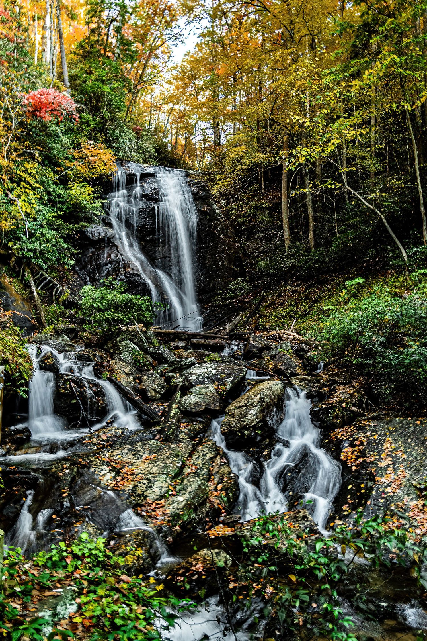Multi-tiered waterfall cascading over rocks surrounded by autumn trees and fallen leaves.
