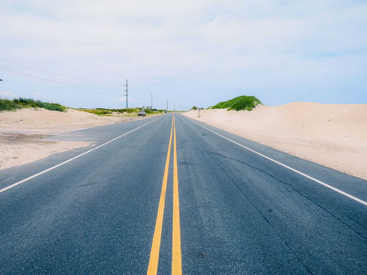 Empty highway stretches toward horizon between sand dunes under bright blue sky.
