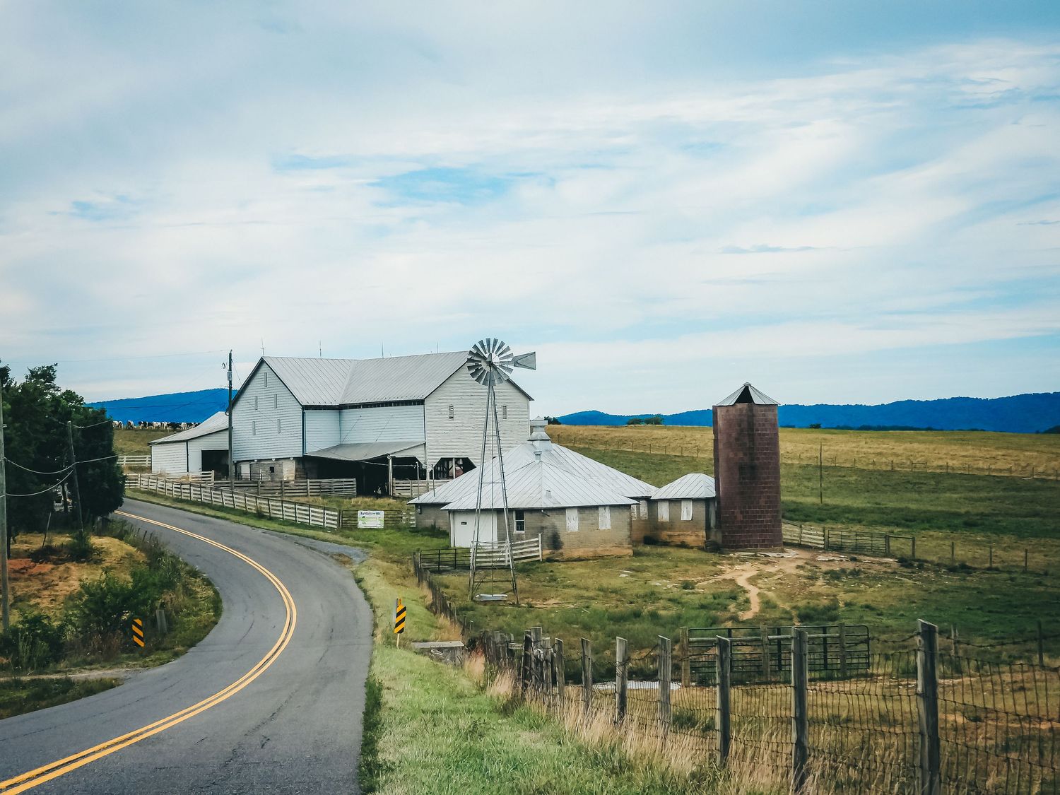 Rural farmland landscape with white barns and silos along a curved country road.