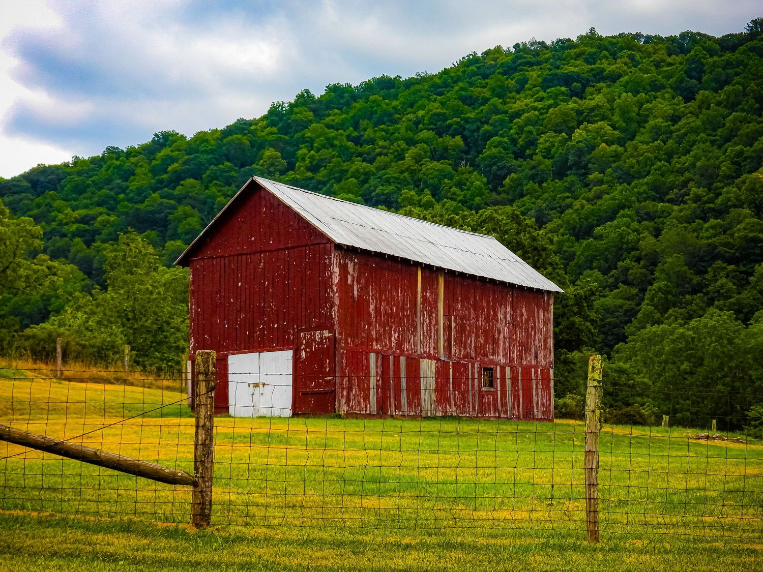 Classic red barn with white trim stands against green hillside behind wire fence.