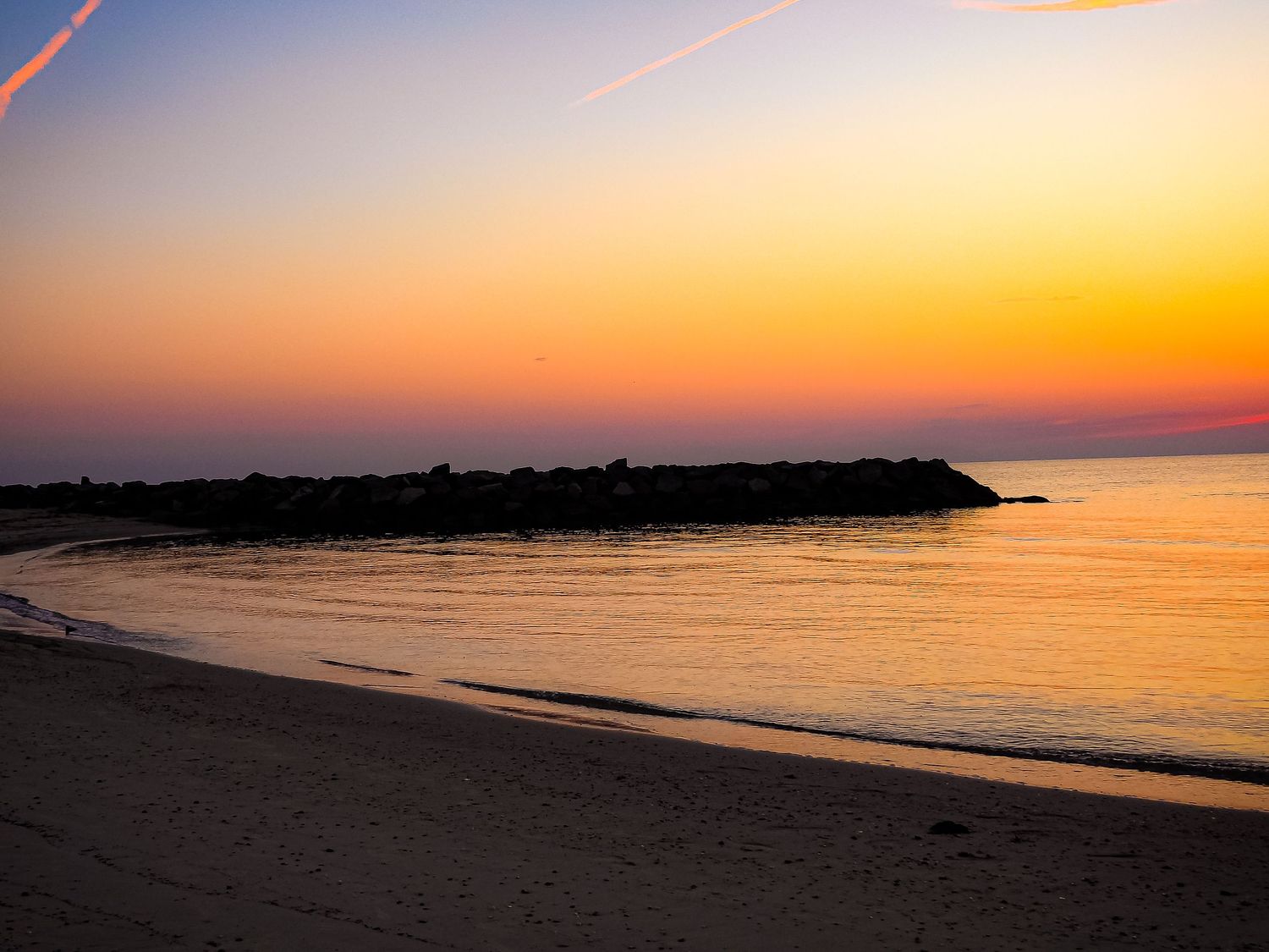Tranquil beach sunset with golden sky reflecting on calm waters near a rocky jetty.
