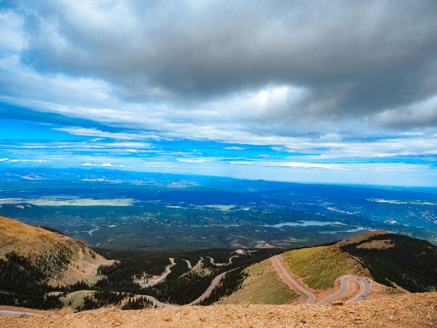 Winding mountain road overlooks a vast valley landscape under dramatic cloudy skies.