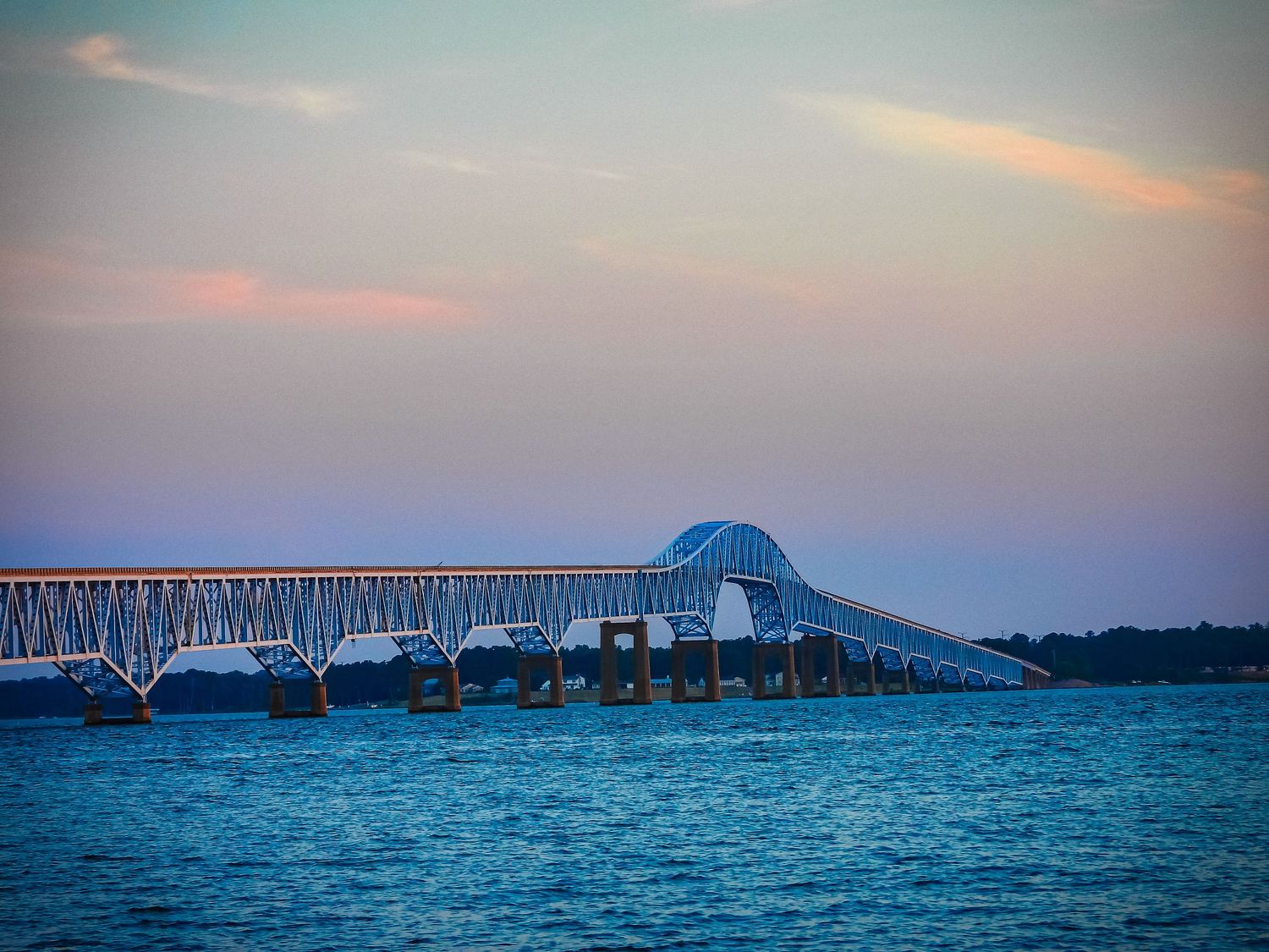 Long blue bridge spans across water during twilight with soft pastel sky.
