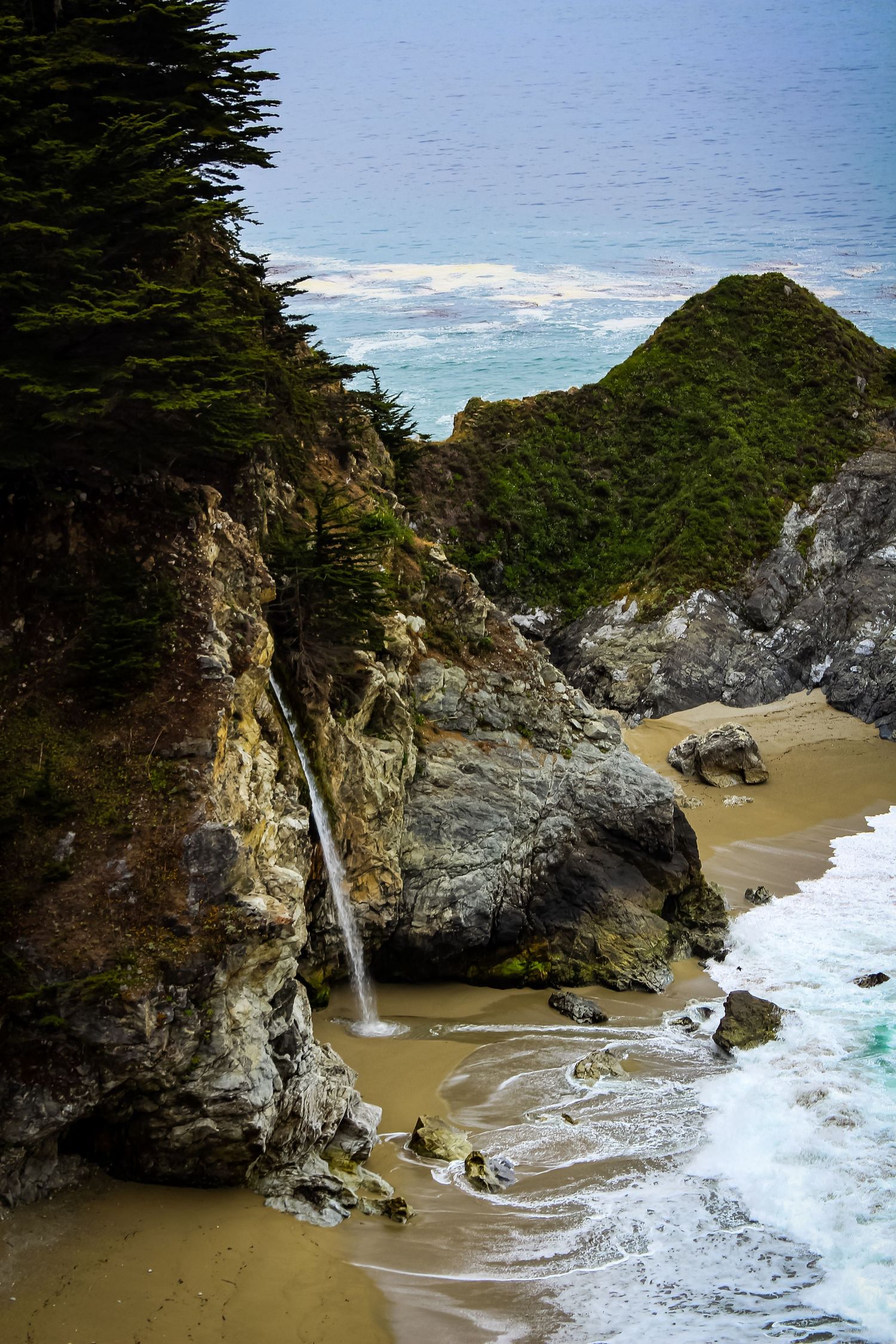 Waterfall cascades onto secluded beach cove with crashing waves and rocky cliffs.