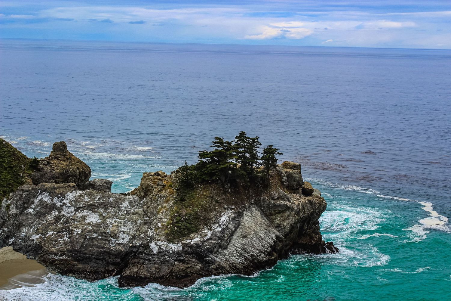 Rocky coastal outcrop with cypress trees surrounded by turquoise ocean waters.