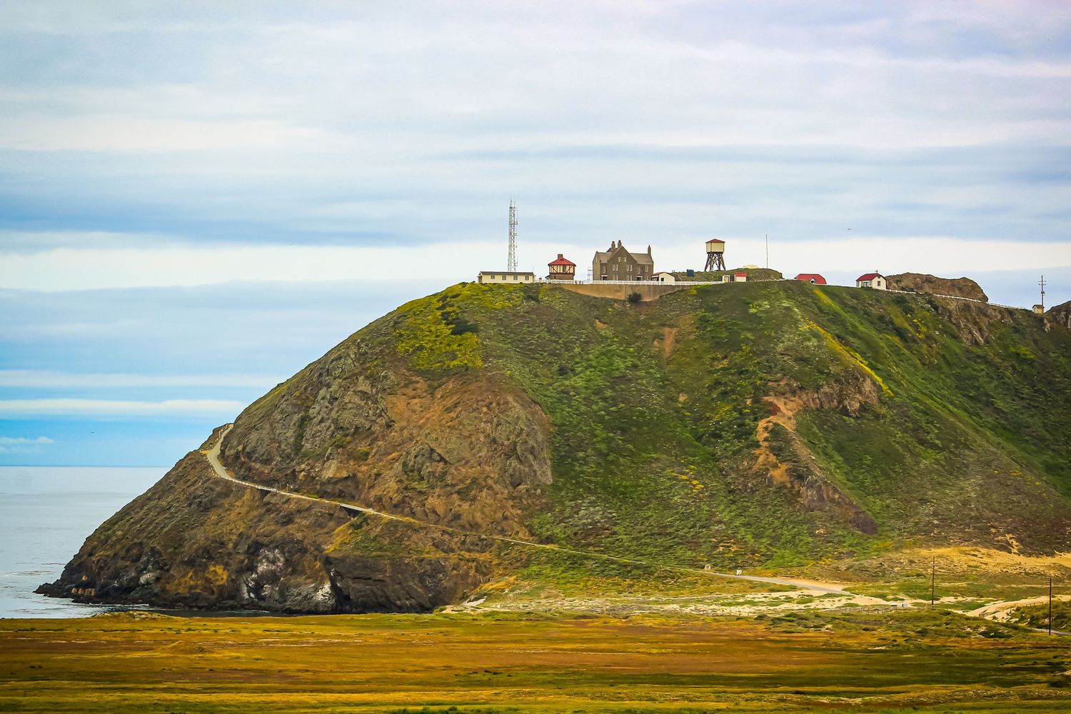 Lighthouse and buildings perched atop coastal hill overlooking Pacific Ocean.