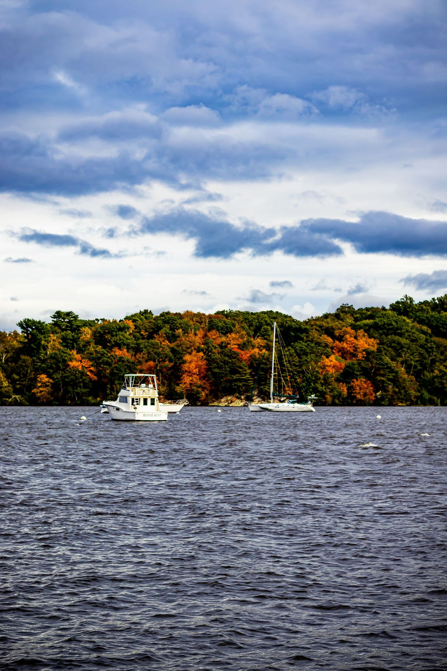 Boats anchored on calm water against vibrant fall foliage and dramatic cloudy sky.
