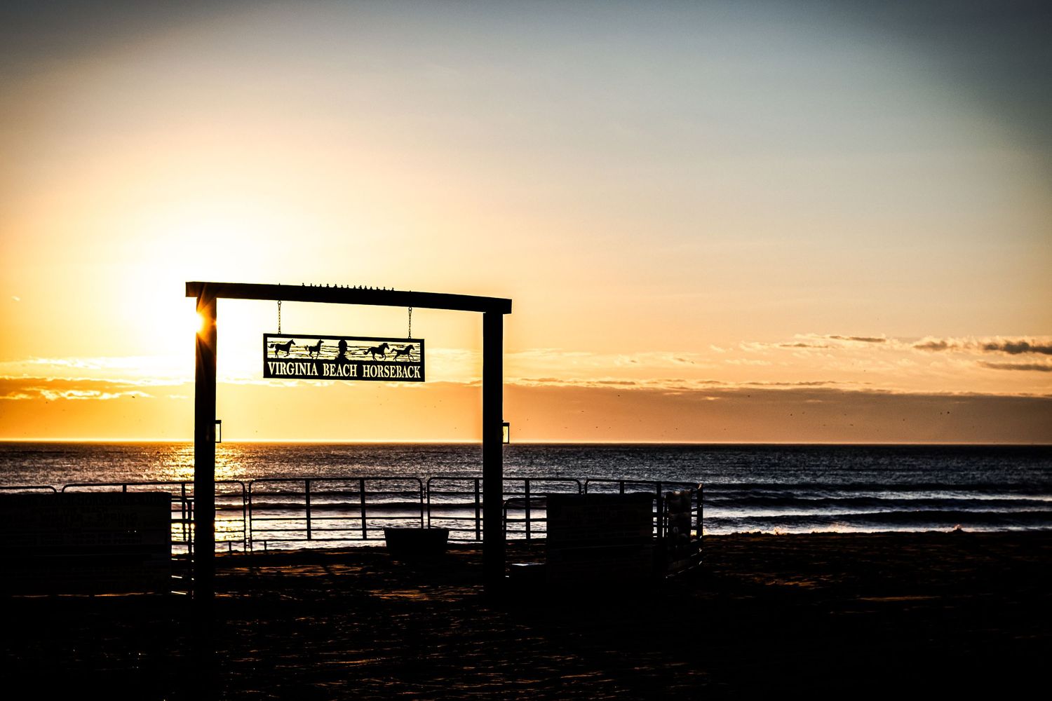 Wooden pier sign silhouetted against a dramatic ocean sunset creating a peaceful coastal scene.