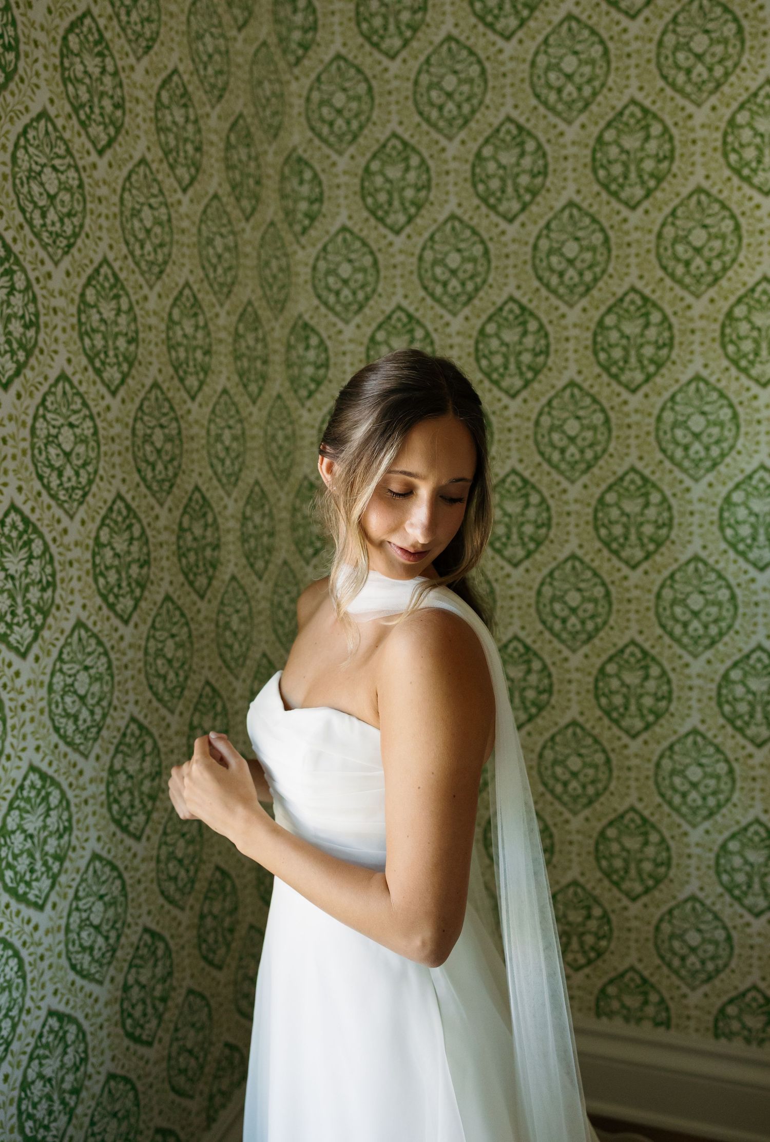 A figure in a flowing white dress poses gracefully against a vintage green damask wallpaper background.