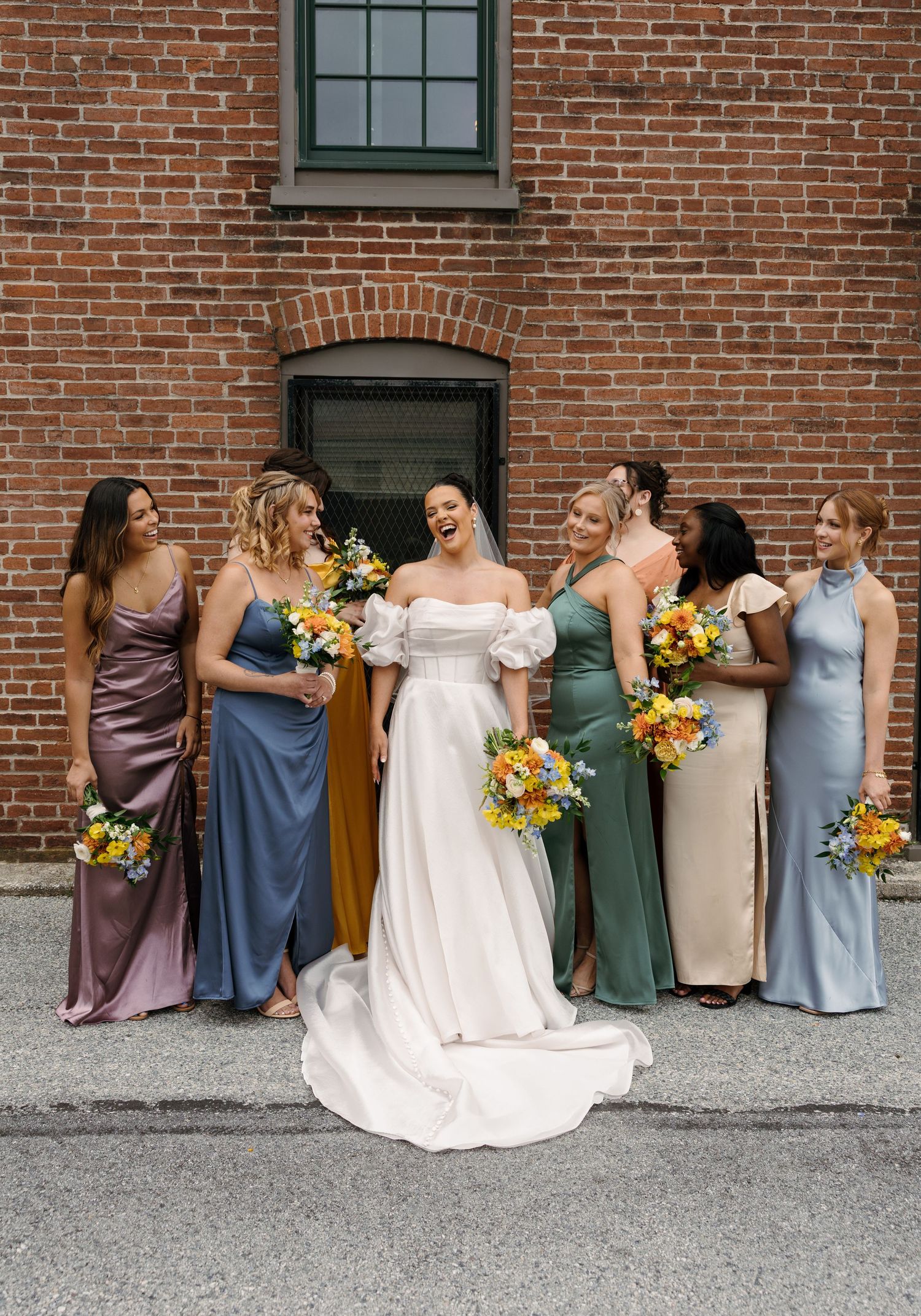 A bridal party in mismatched dresses of purple, blue, green and beige holds yellow sunflower bouquets against a brick wall.