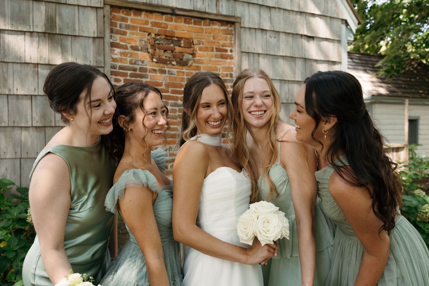 Bridesmaids in sage green dresses share joyful moments during wedding photos against a brick wall.