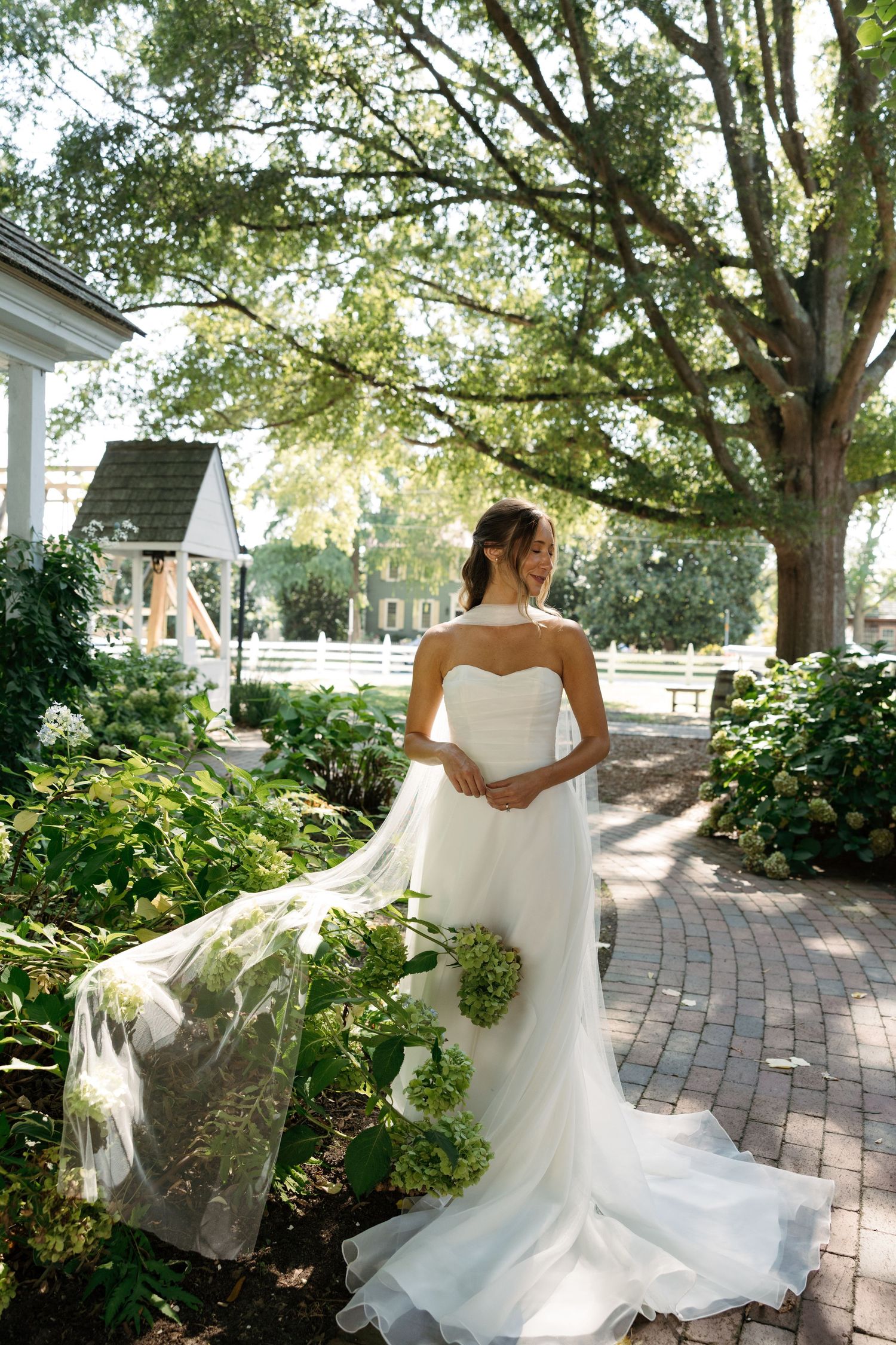 A flowing white wedding dress twirls gracefully on a brick pathway surrounded by lush greenery and trees.