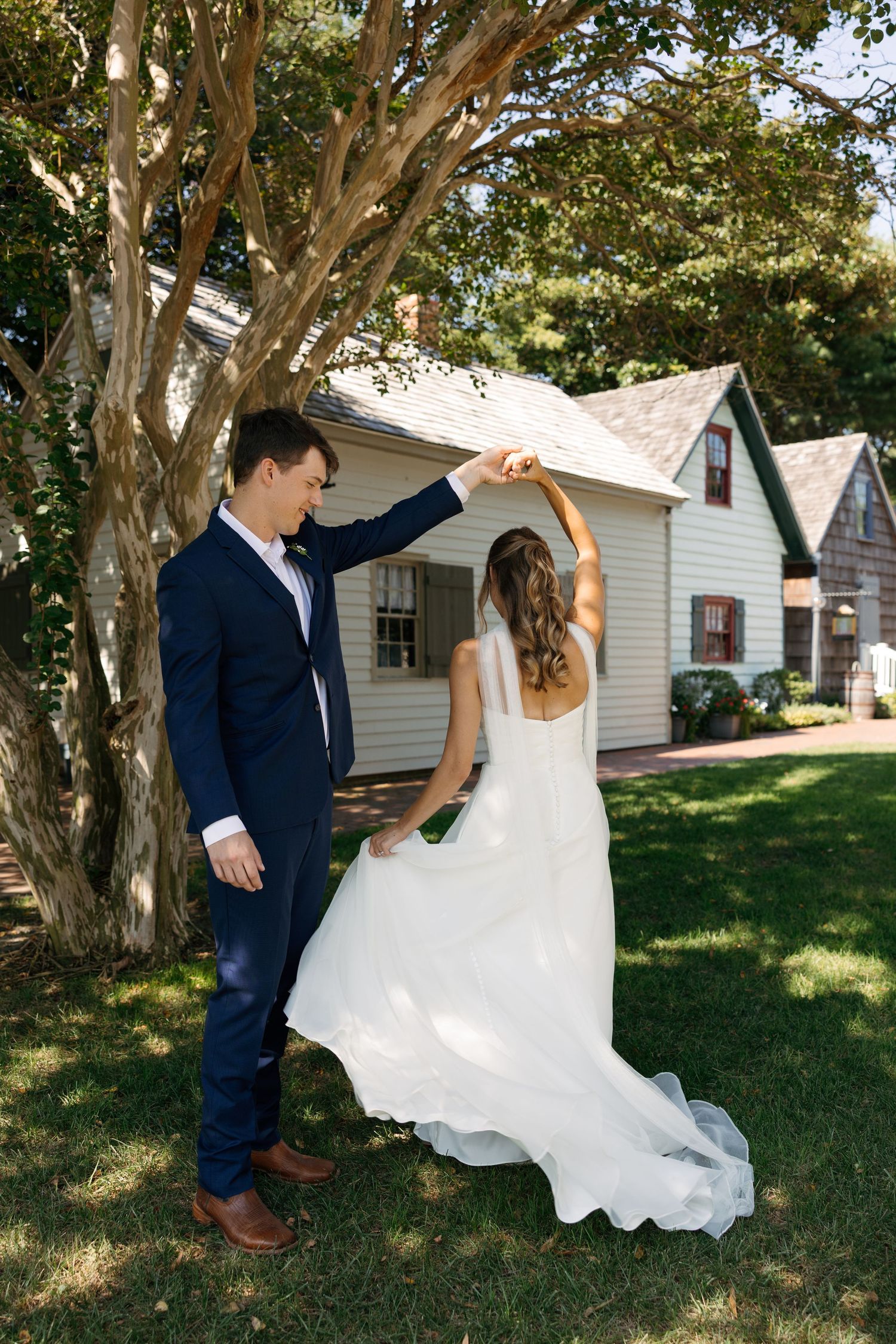 A couple in wedding attire dances and twirls together on a sunny lawn with white cottages in the background.