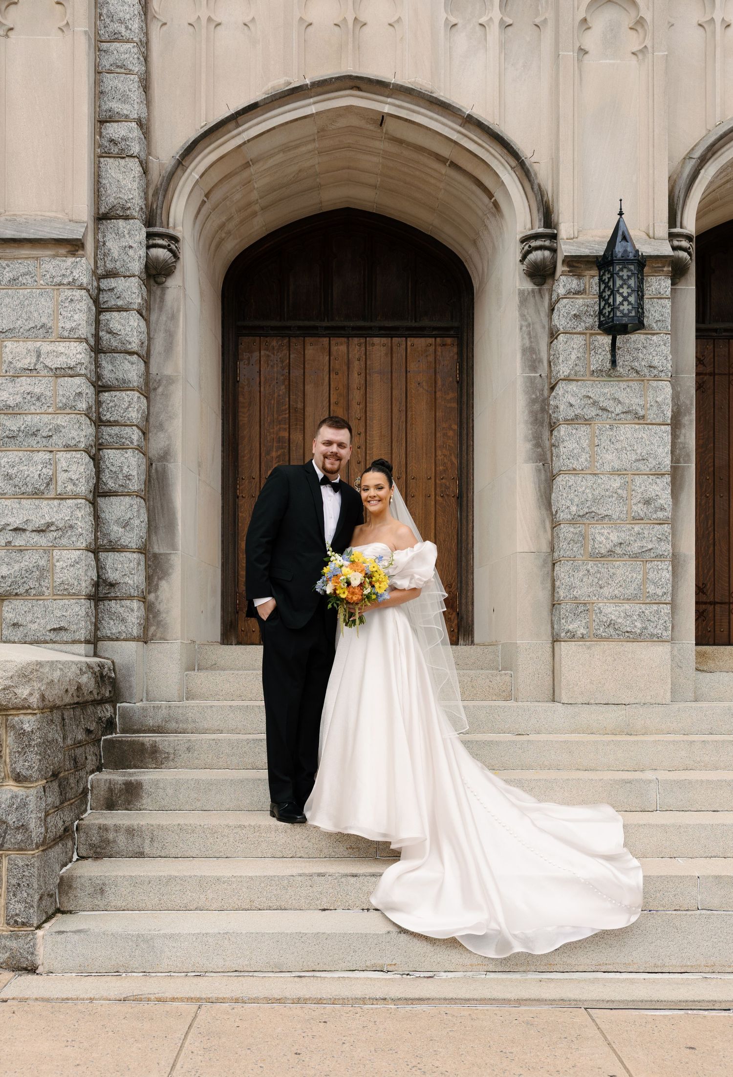 A couple in wedding attire poses on stone church steps in front of an arched wooden doorway with a lantern.