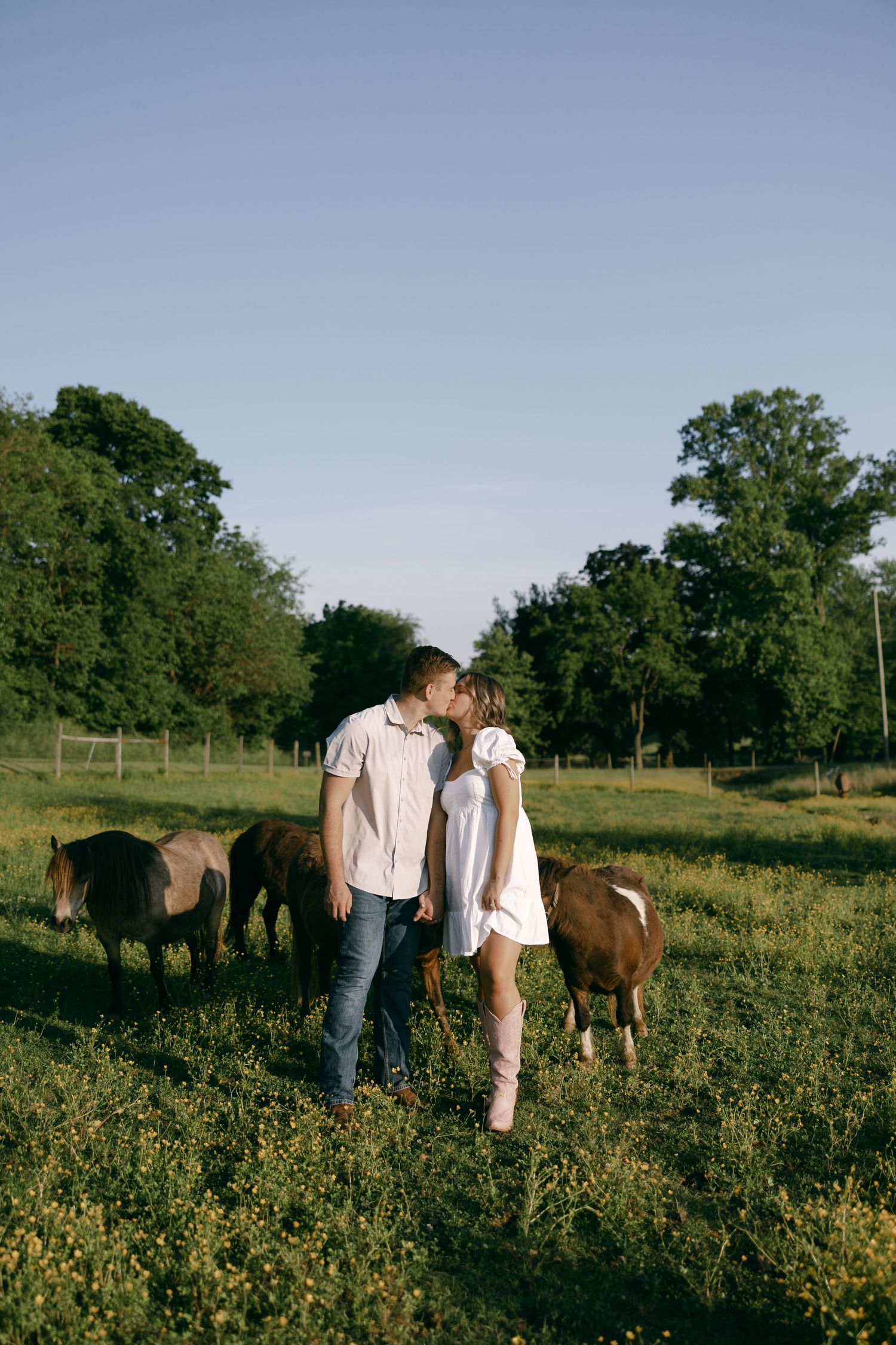 Couple sharing a romantic moment in a field with horses grazing in the background.