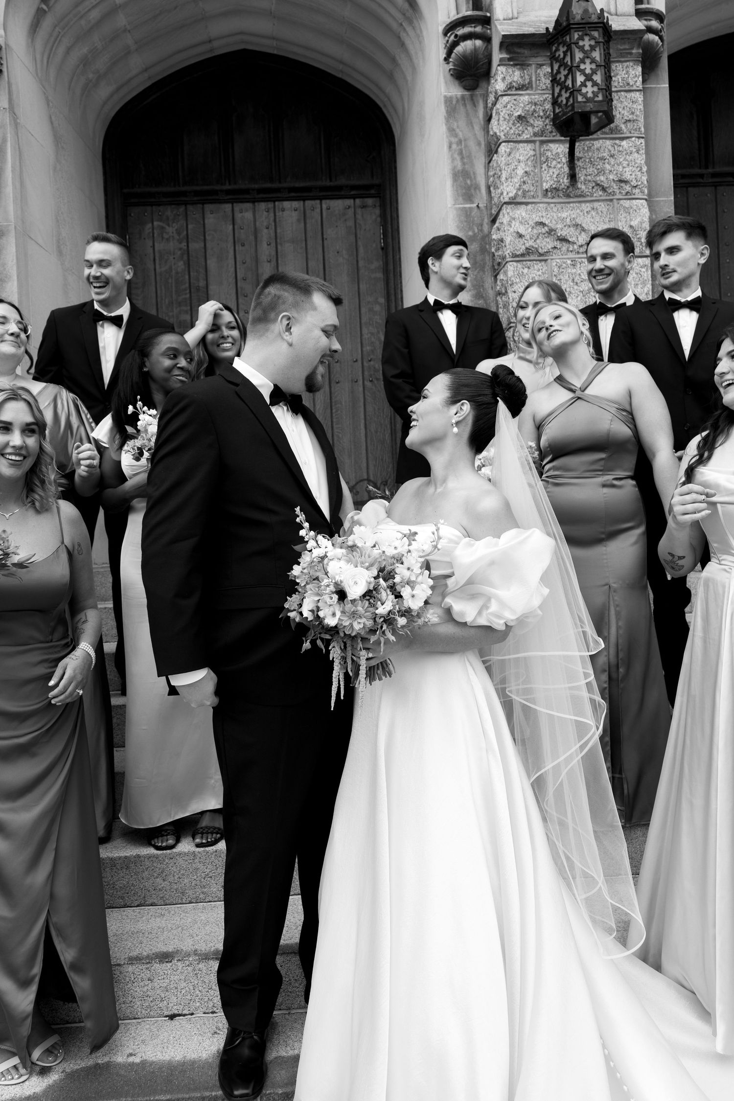 Wedding party gathers outside church entrance with bride in white dress holding pink and white floral bouquet.