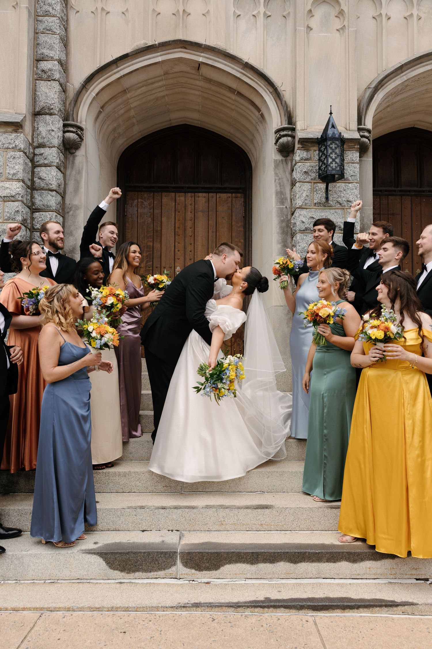 Wedding party in colorful dresses of yellow and blue poses on stone steps with arched doorways in background.