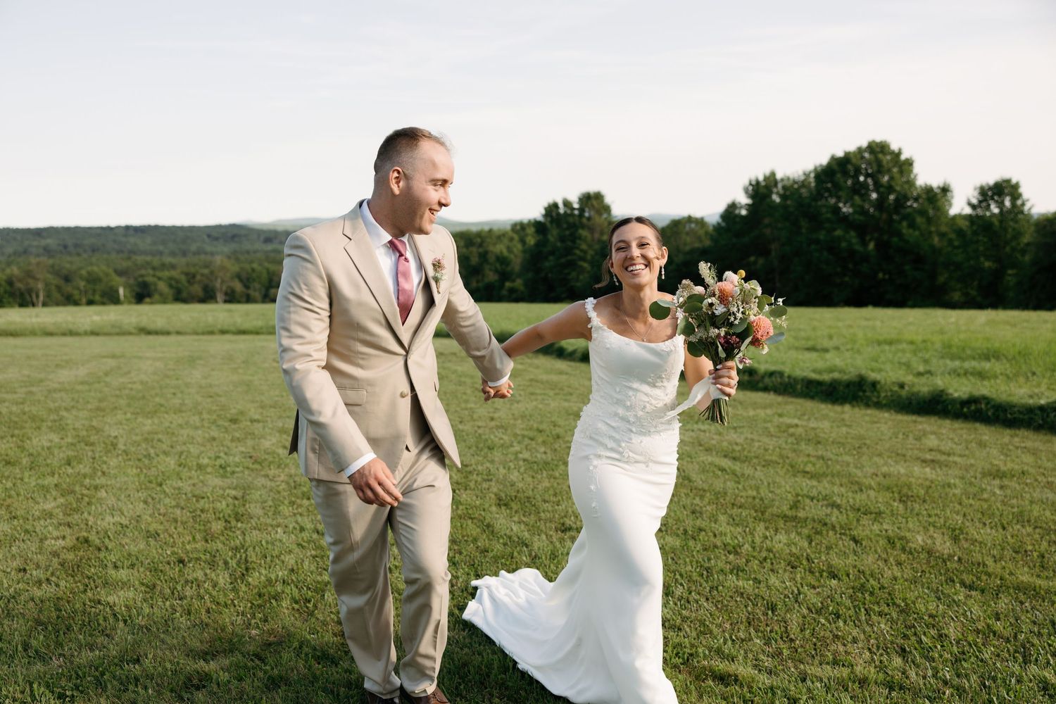 A couple in wedding attire dances and celebrates joyfully in a sunny green meadow.