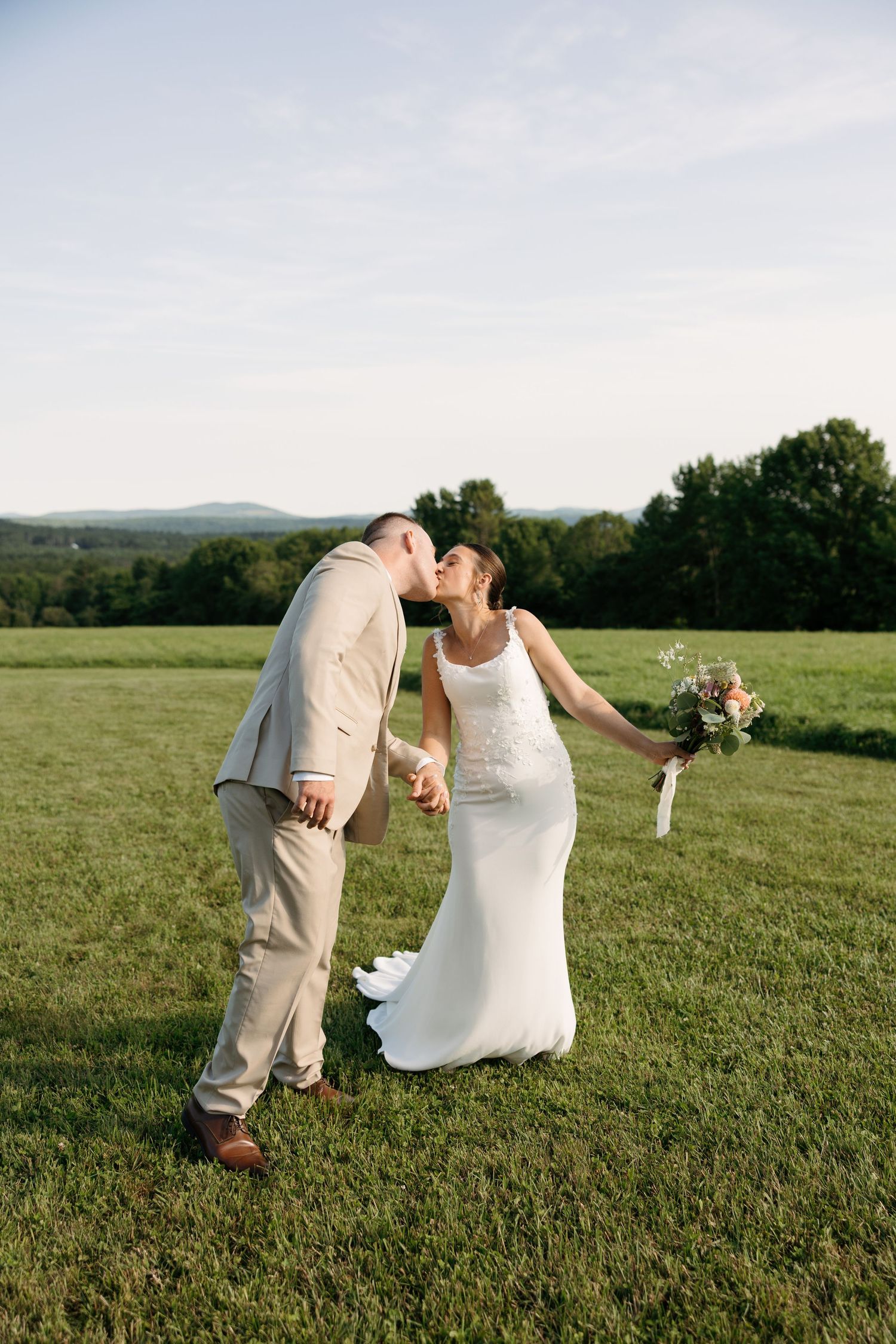 A couple in wedding attire playfully dips and poses together on a grassy hilltop with scenic views.