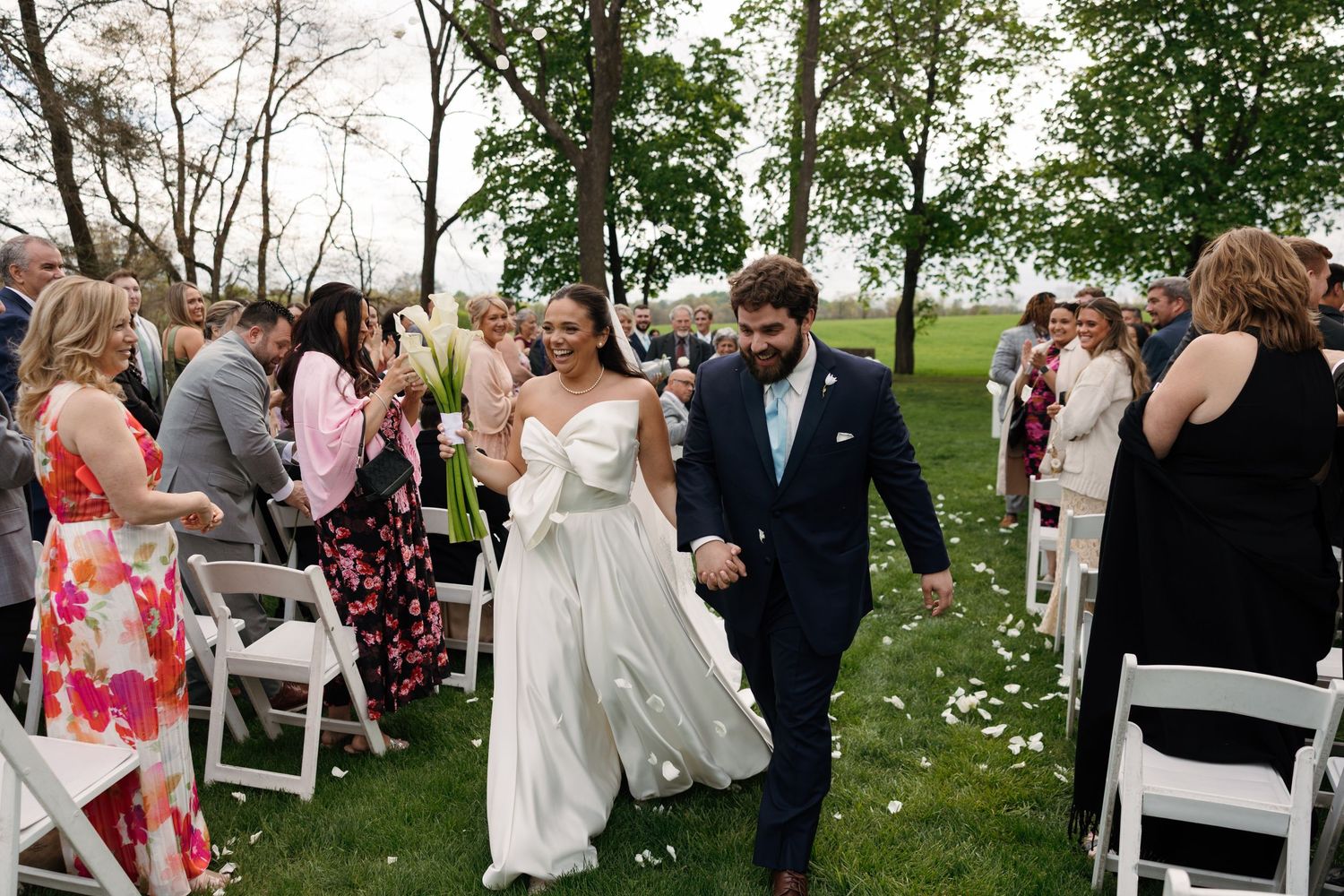 Bride and groom walk down outdoor wedding aisle holding hands, guests cheering, white chairs, green trees backdrop.