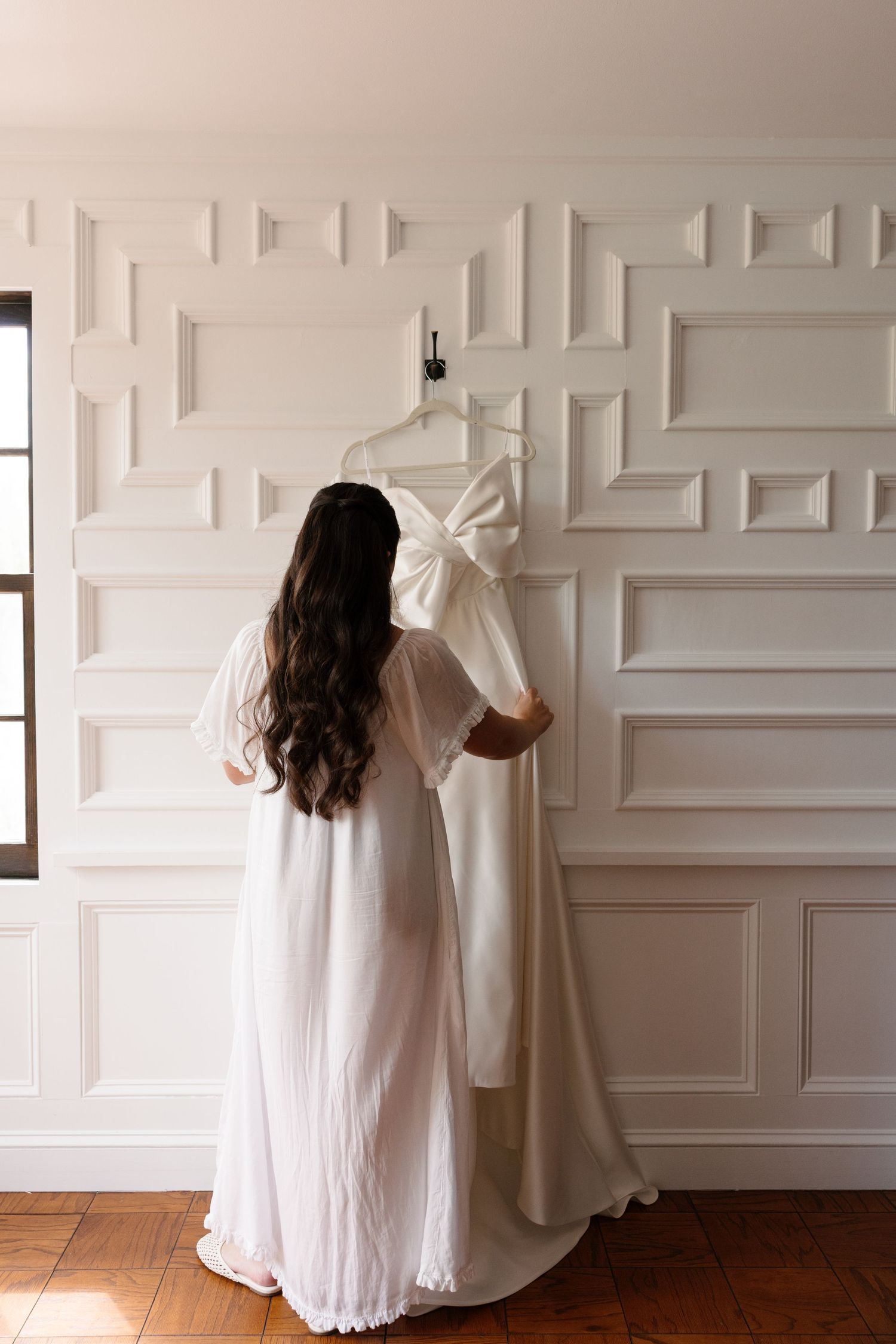 Bride in white robe admiring her wedding gown hanging on elegant white paneled wall in a bright room.