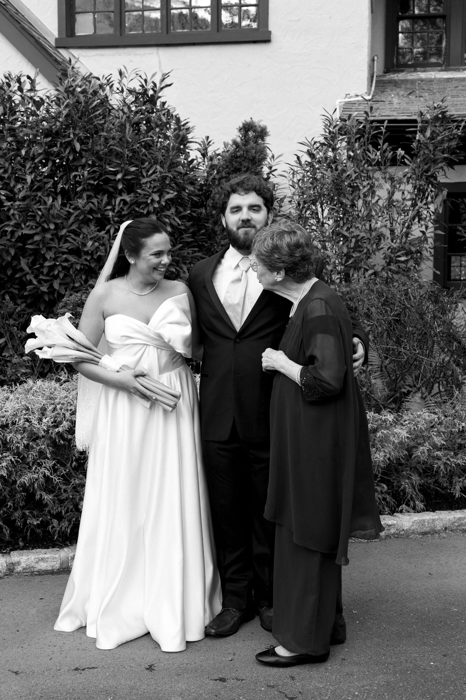 Black and white wedding photo of bride in white gown, groom in tuxedo, and older woman posing together outdoors.