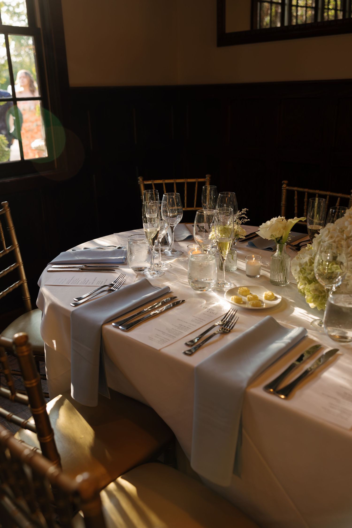 Elegant round dining table set with white linen, silverware, wine glasses, and floral centerpieces in a dimly lit room.