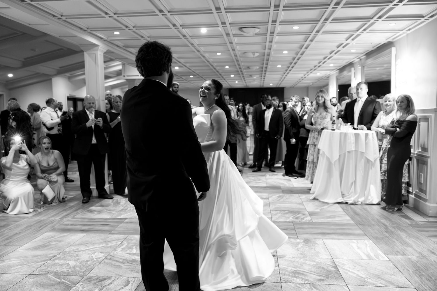 Black and white photo of bride and groom sharing first dance at elegant wedding reception with guests watching.