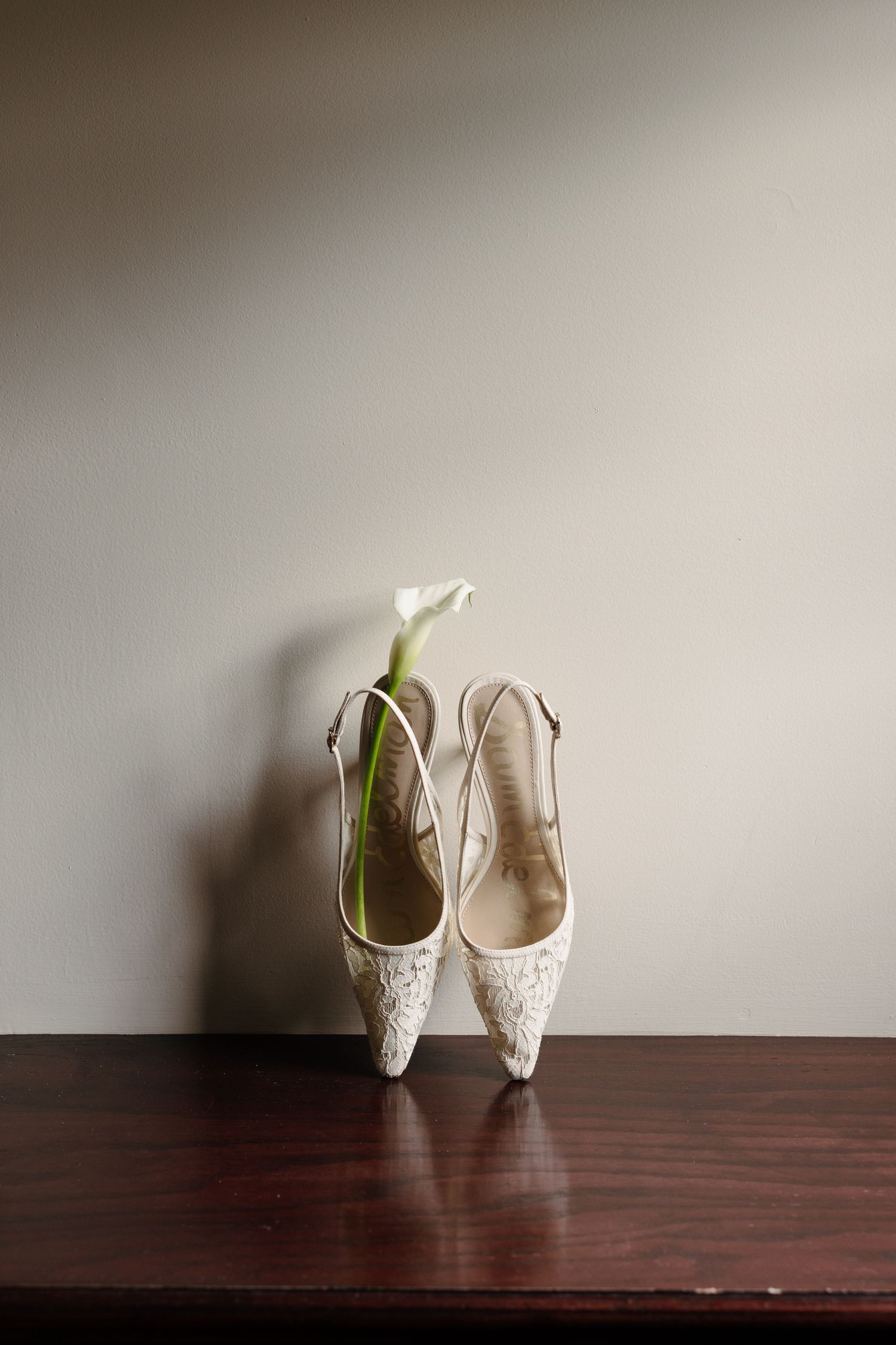 White lace bridal slingback heels with a calla lily flower on a dark wood surface against a neutral wall.
