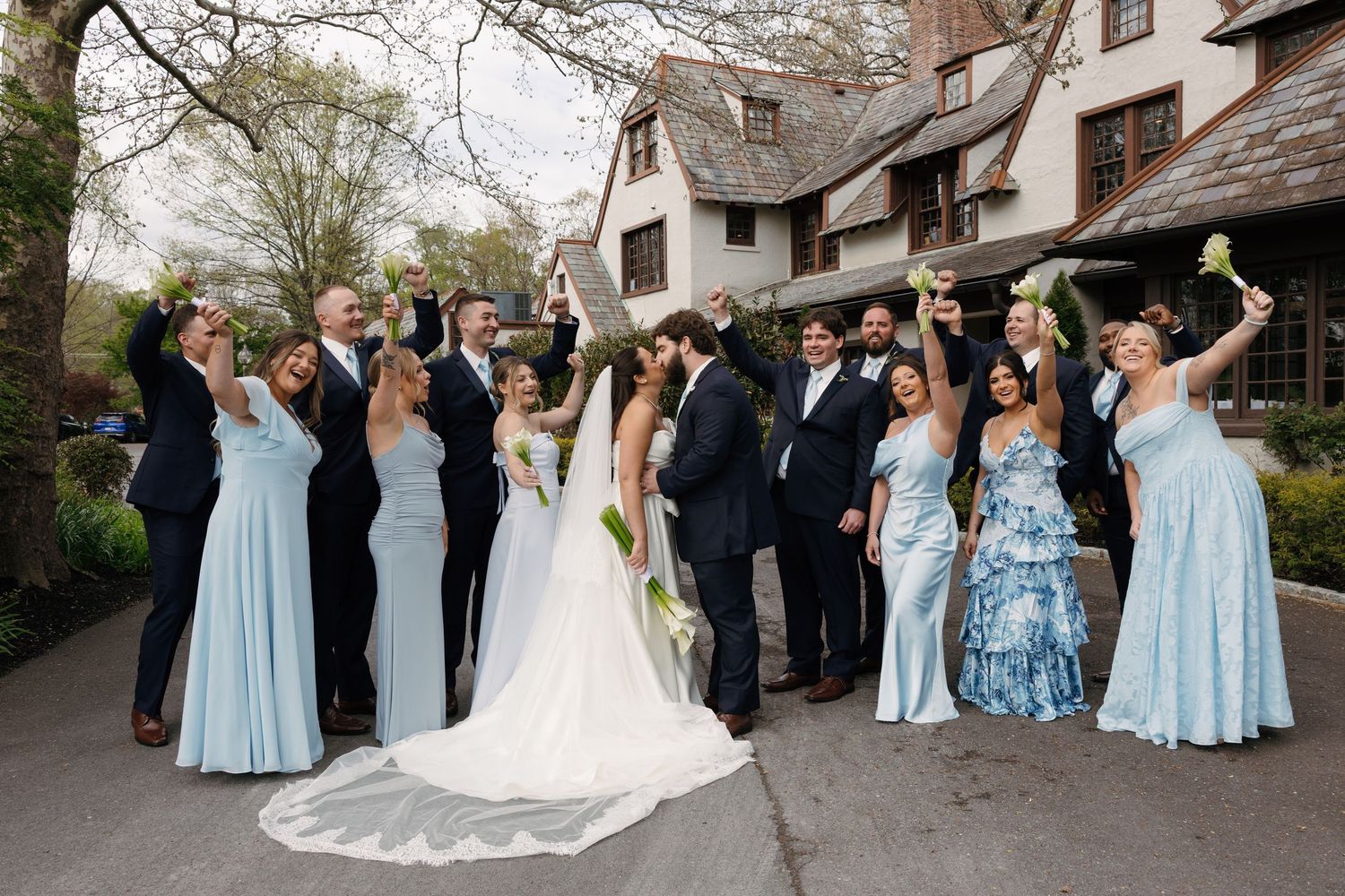 Wedding party celebrating outdoors in light blue bridesmaid dresses and navy suits, bride in white gown, raising glasses.