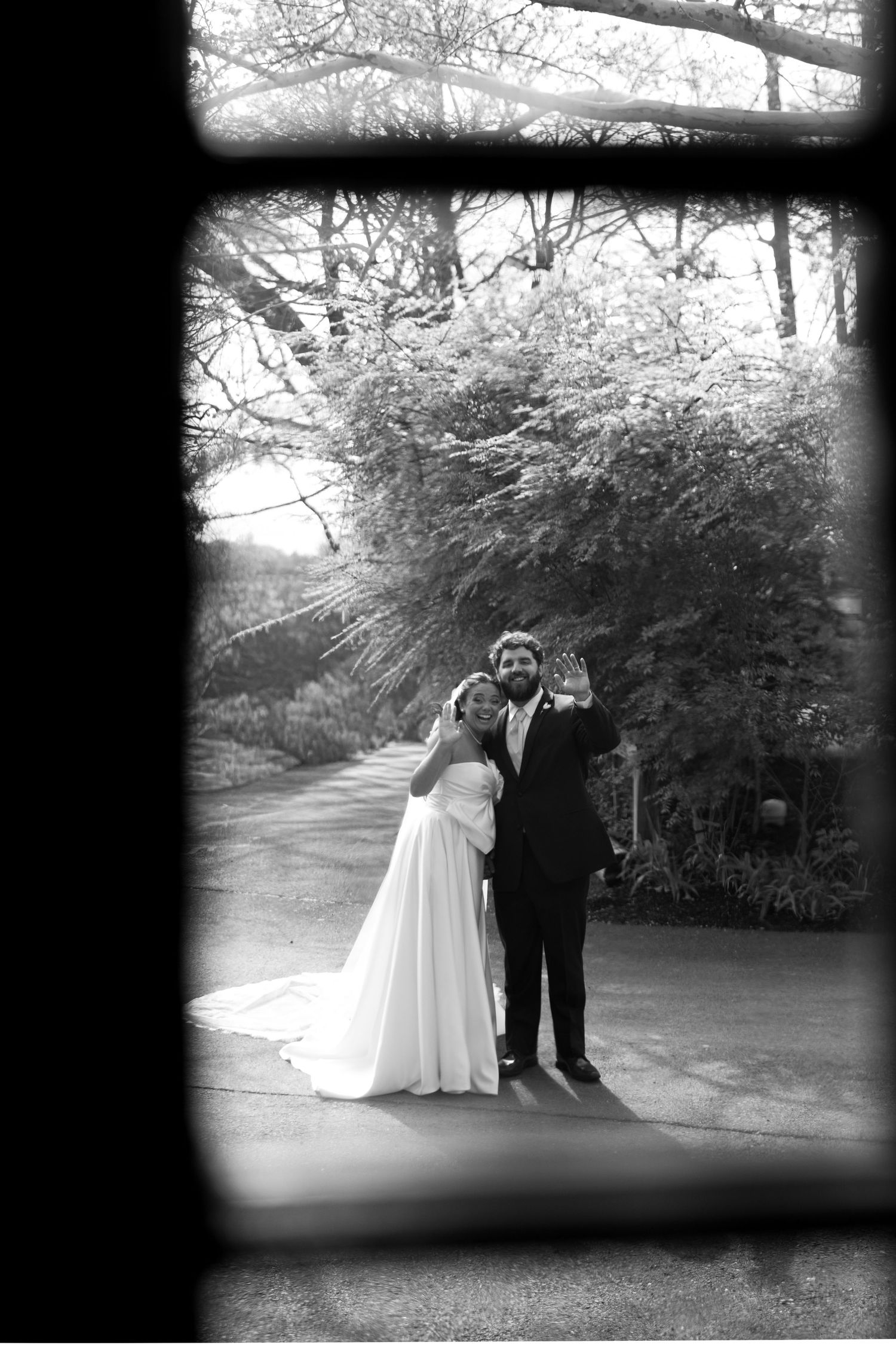 Black and white wedding photo of bride and groom posing outdoors, framed by a window, surrounded by lush trees.