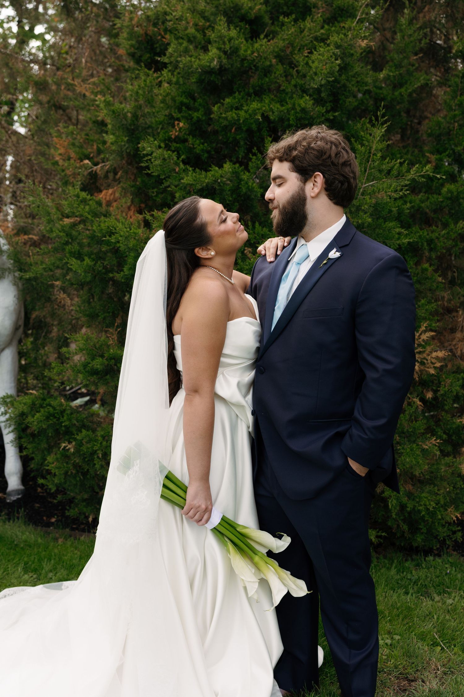 A bride in a white gown holding calla lilies gazes at her groom in a navy suit outdoors near evergreen trees.