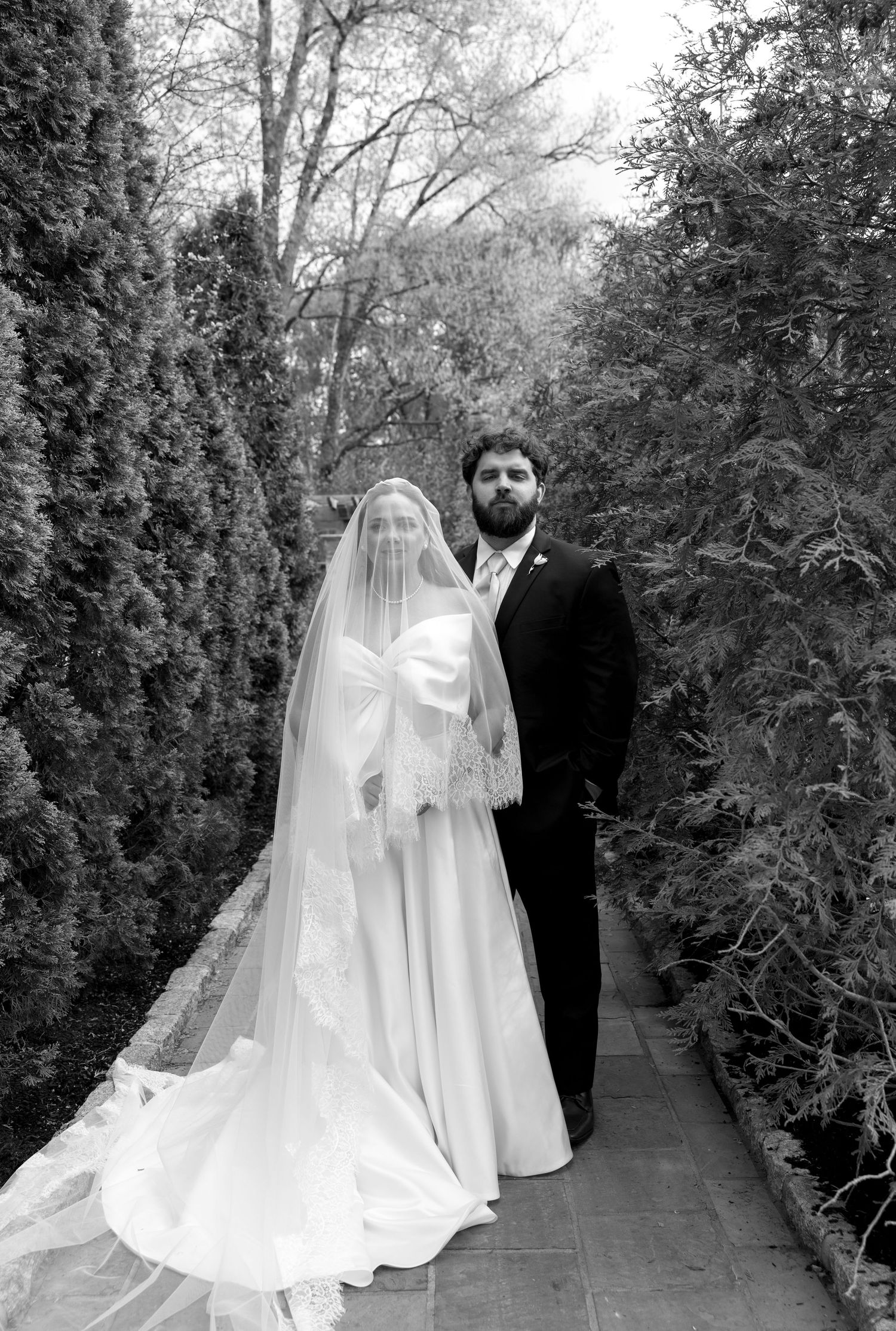 Black and white wedding photo of bride in veil and gown with groom in tuxedo standing in garden pathway.