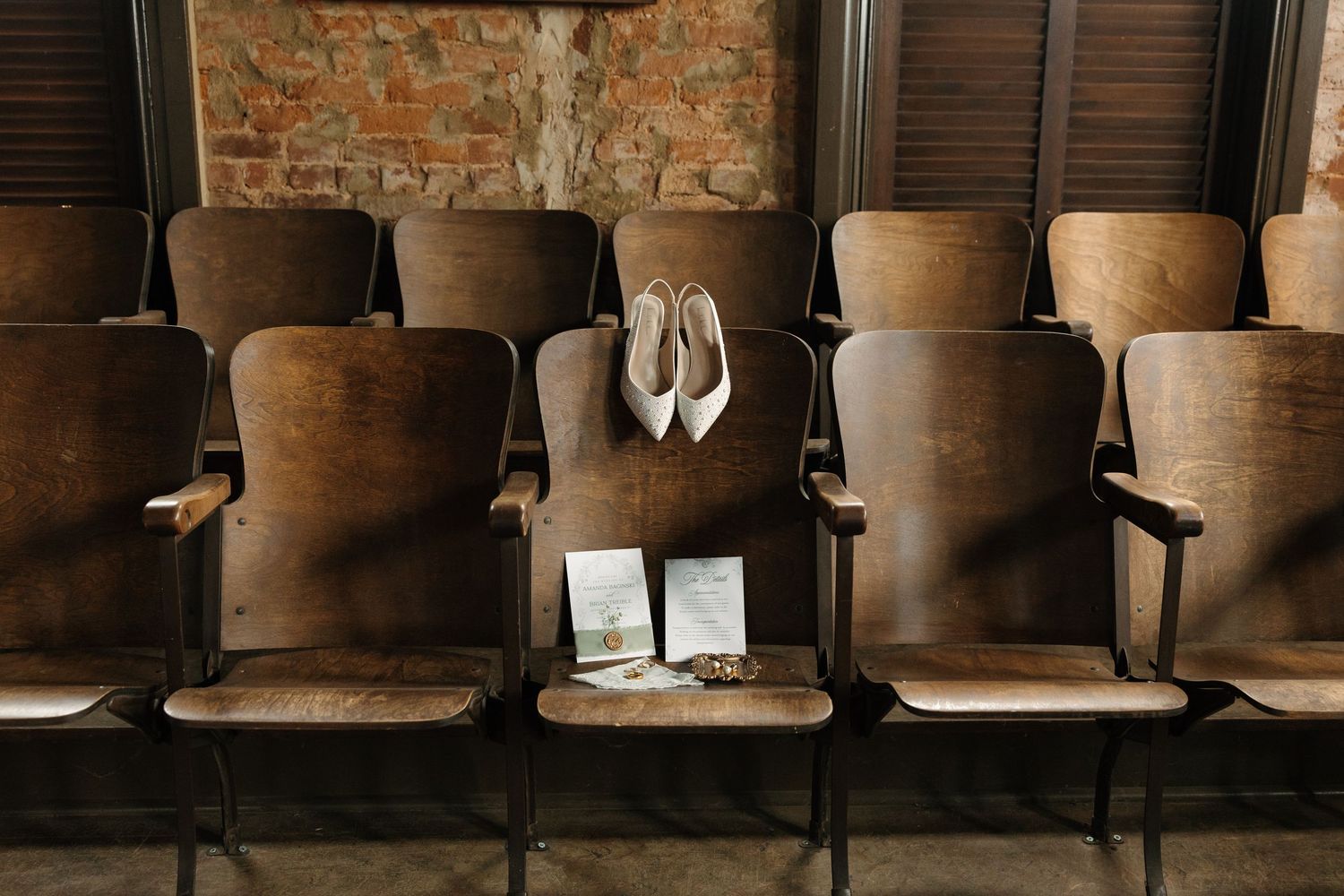A row of vintage wooden theater seats with red velvet upholstery lines up against a rustic brick wall.