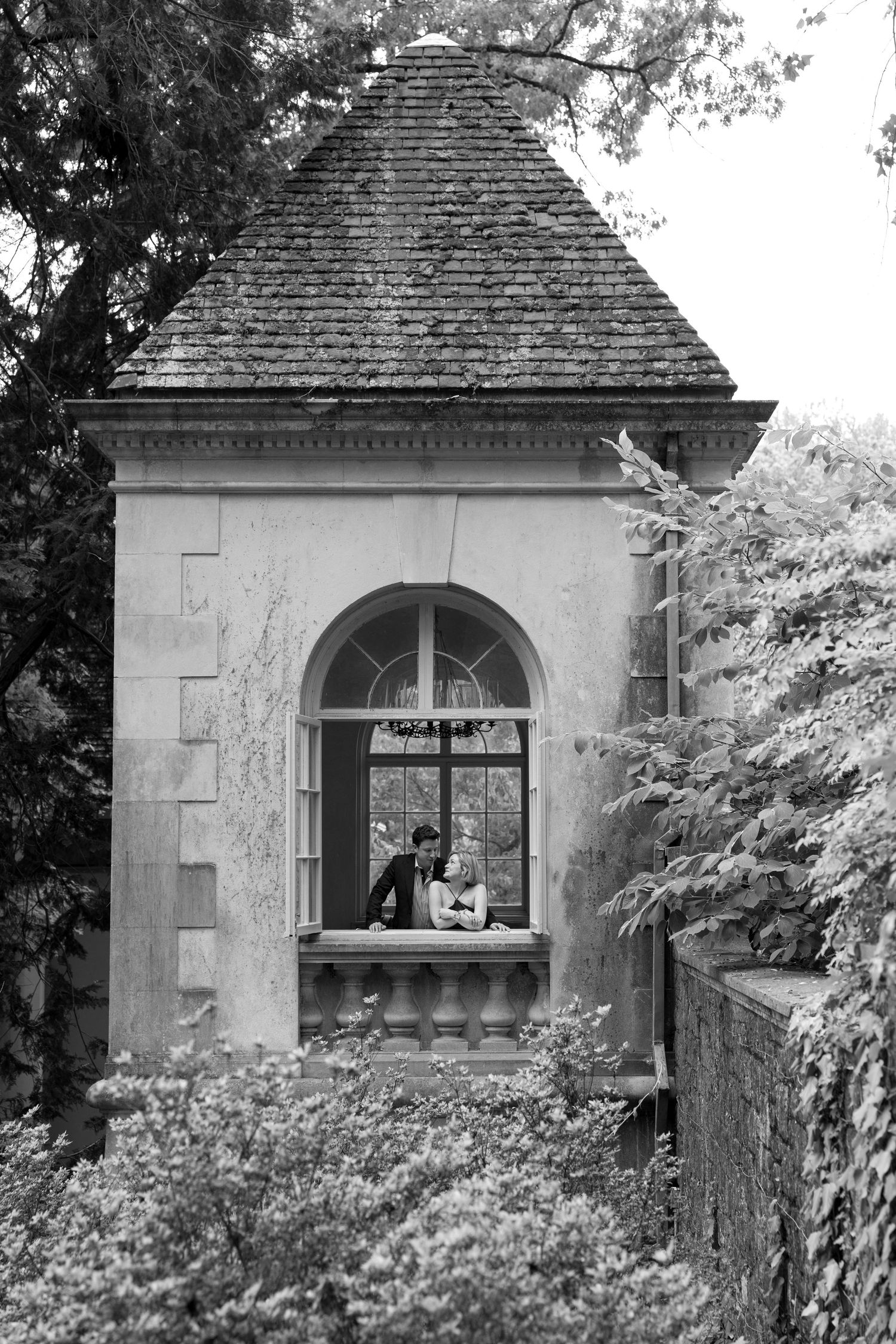 Black and white photo of couple sitting in arched window of elegant stone garden pavilion surrounded by trees.